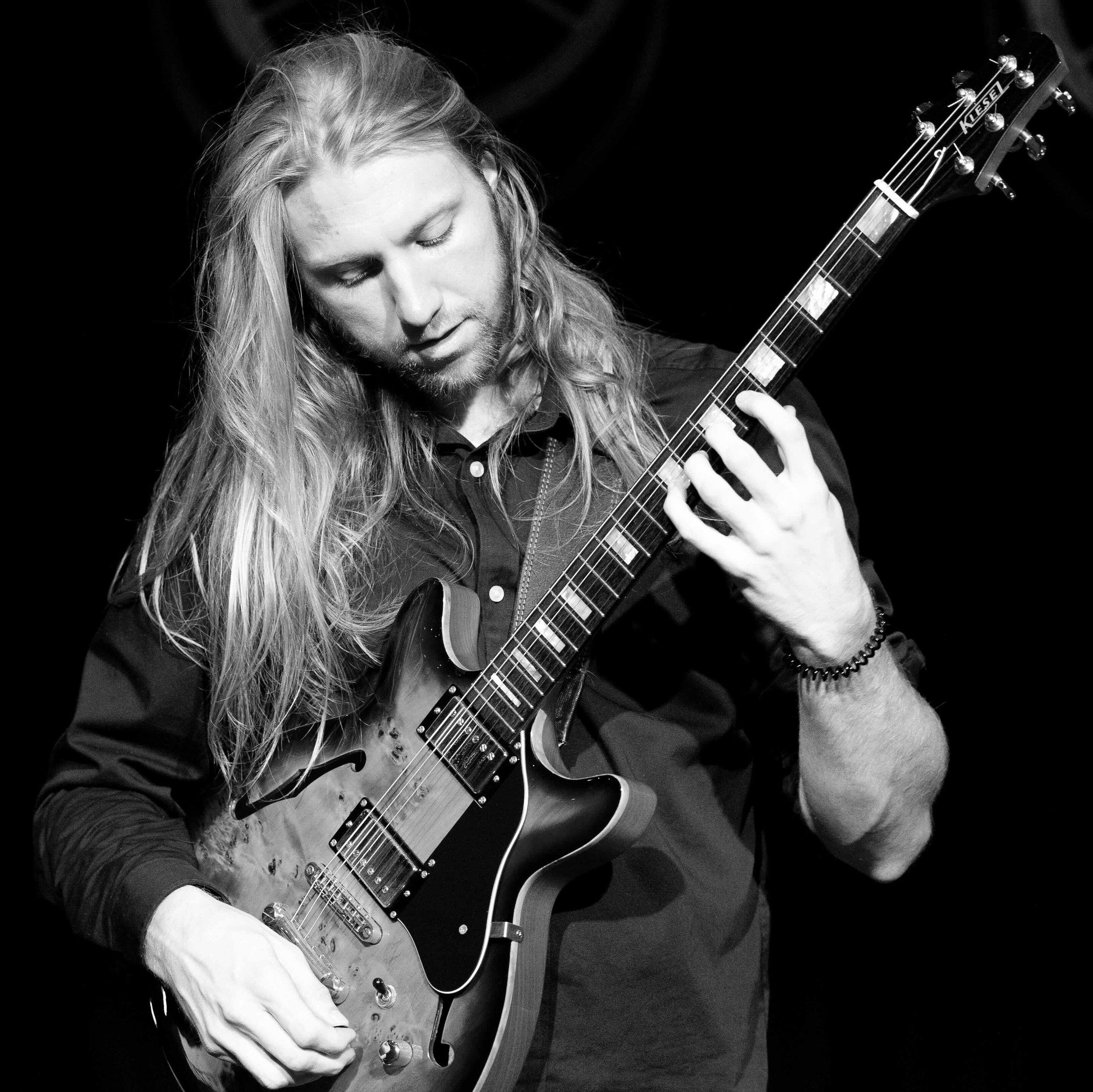 A man with long hair and a beard playing an electric guitar with closed eyes.