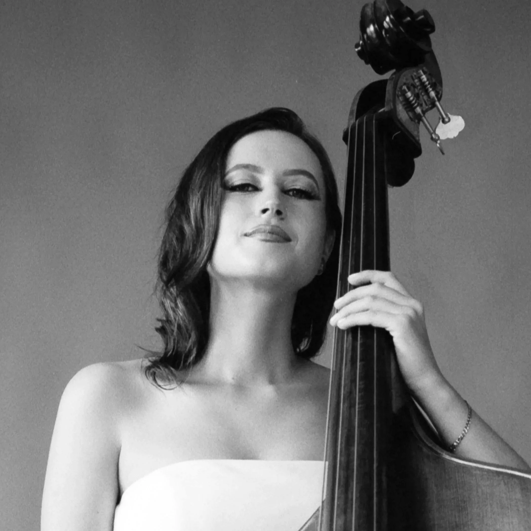 A black and white photo of a woman with long hair, partially visible, holding a violin by the neck, with a confident expression, against a plain background.