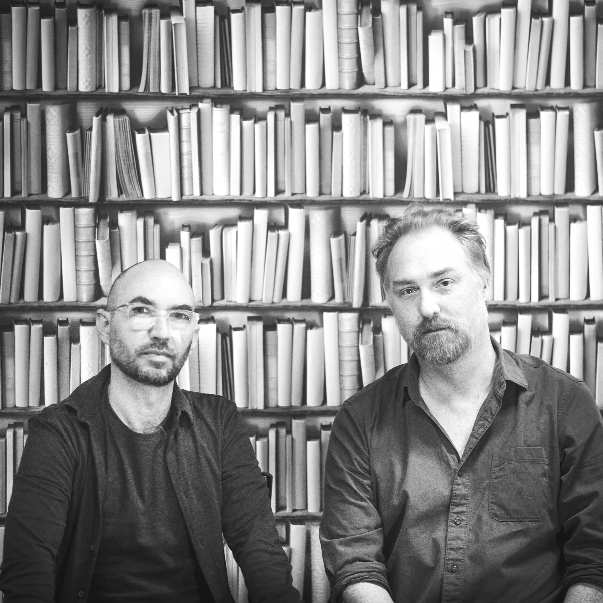 Two jazz musicians with glasses and a beard and a man with short hair and a mustache pose in front of a bookshelf filled with books.