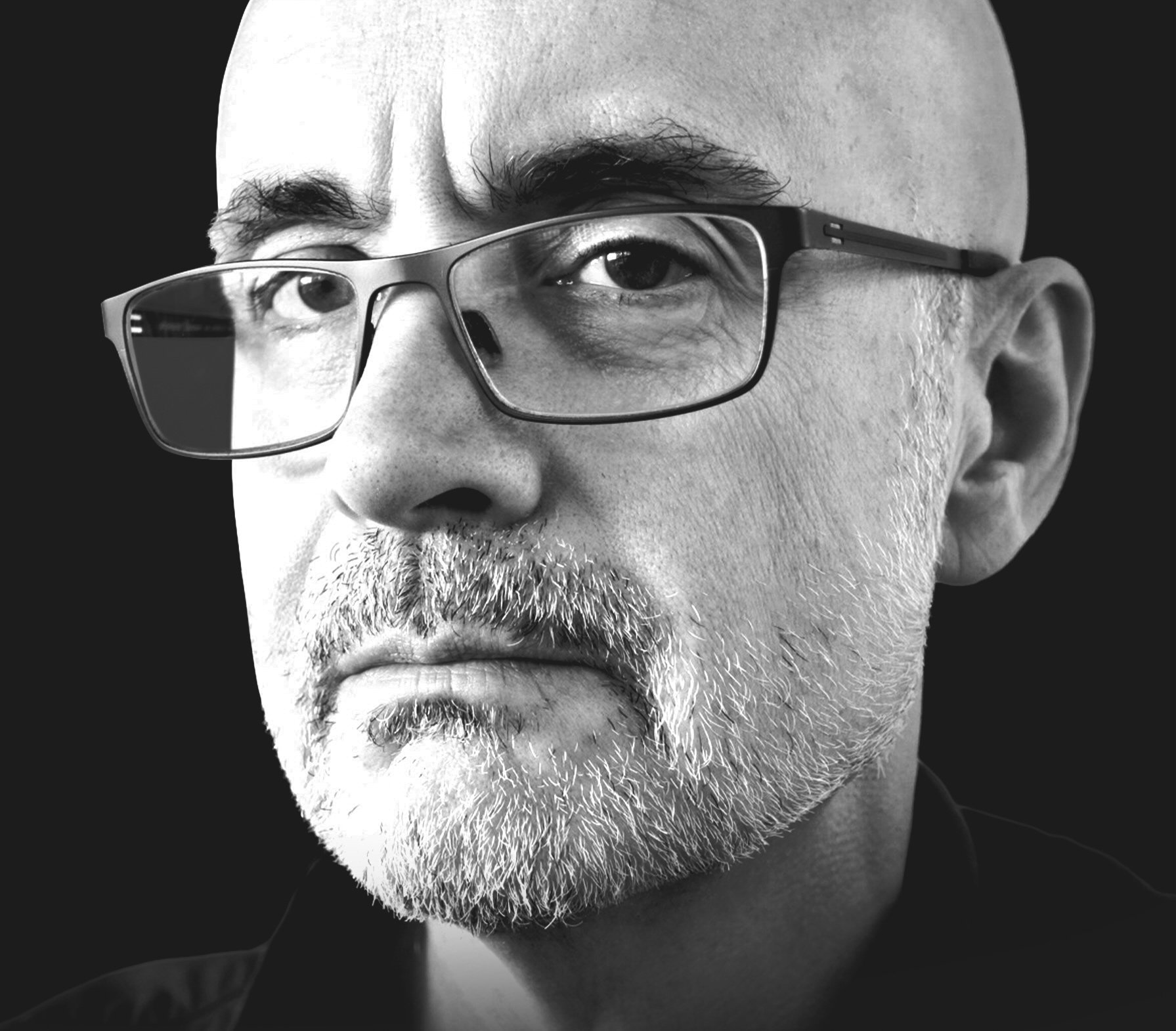 Close-up black and white portrait of a man with a bald head, wearing glasses, and a beard, looking directly at the camera.