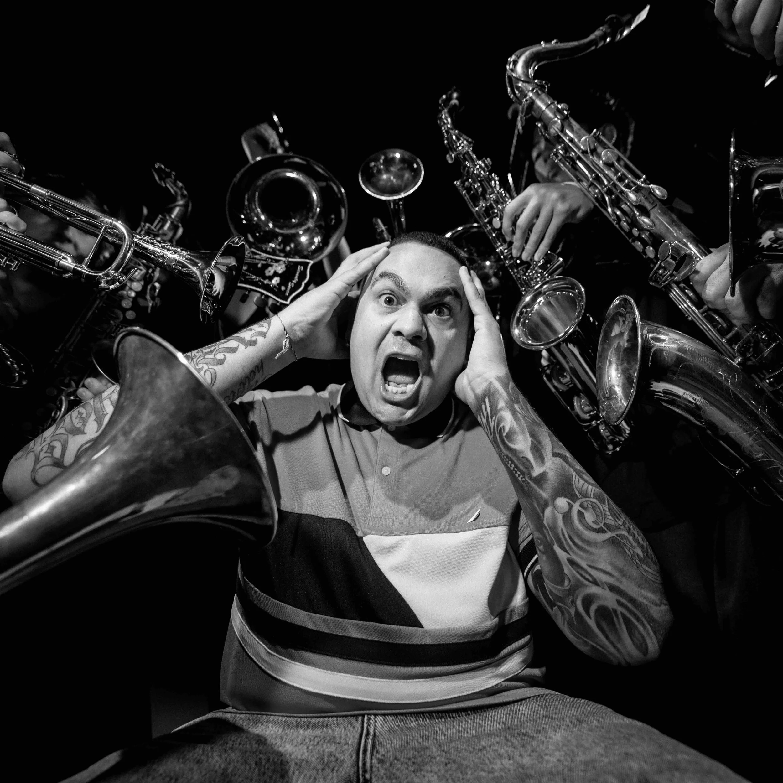 A man with tattoos on his arms looks upward with surprise or excitement as he is surrounded by musical instruments, mainly saxophones.