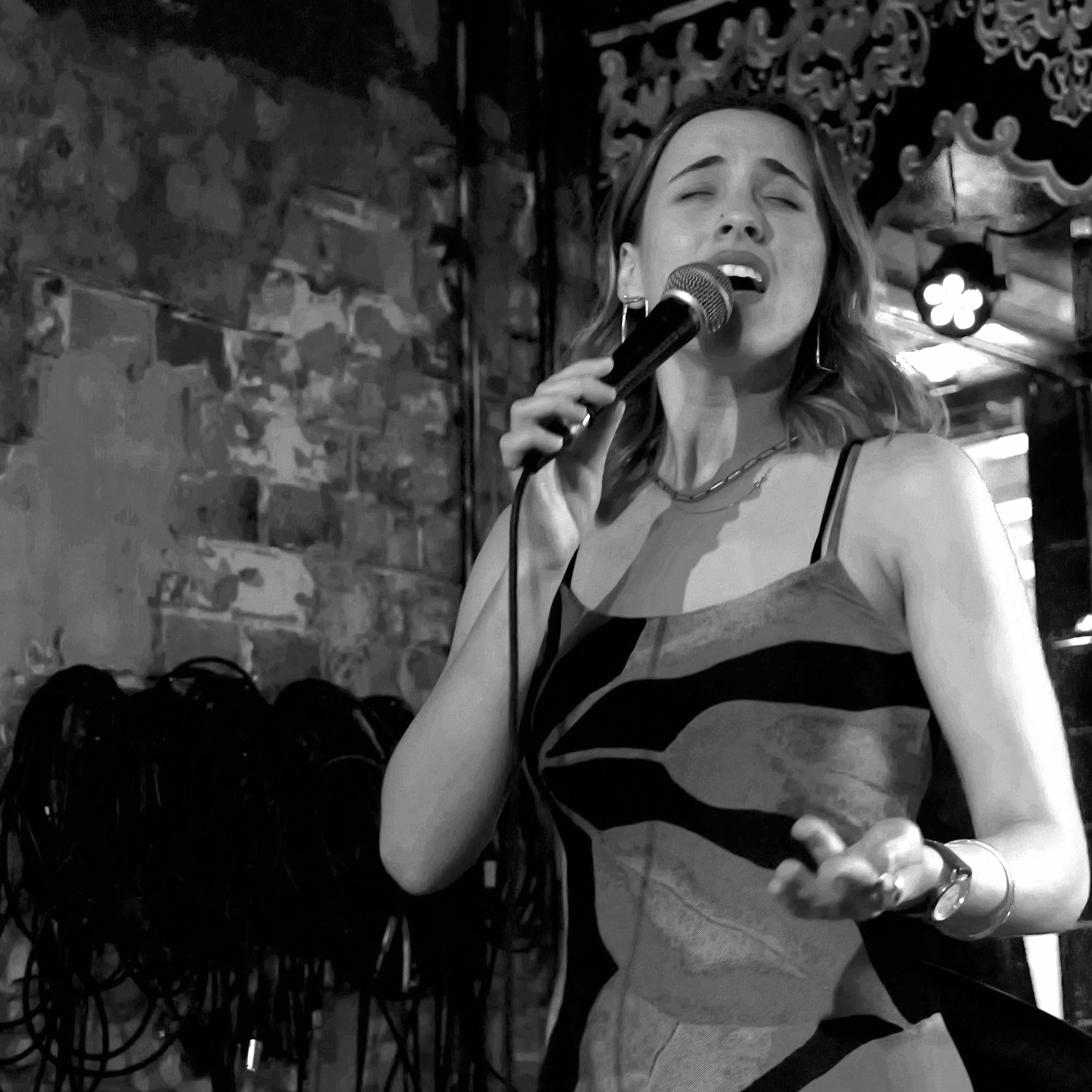A woman singing into a microphone at a bar or club with ornate dark wood decor in the background.