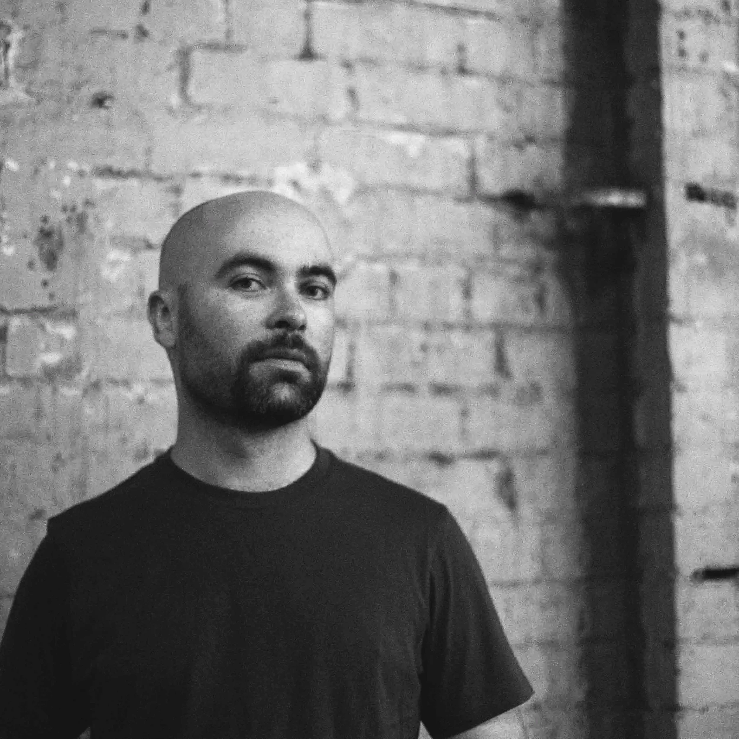 Black and white photo of a man's face, showing only the upper part of his face, forehead, eyes, nose, and beard, set against a textured brick wall background.