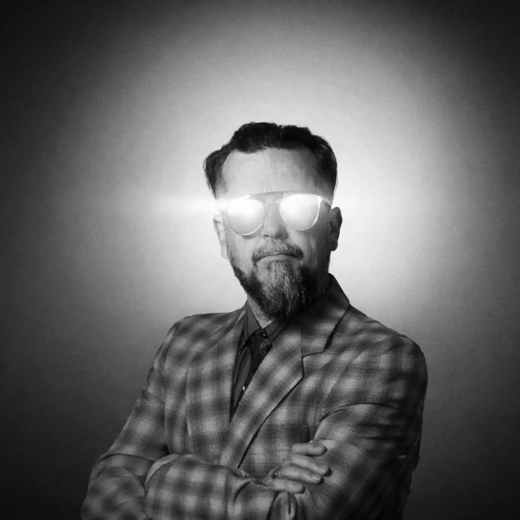 Black and white portrait of a man with a beard and styled hair wearing sunglasses, a checked blazer, and a shirt, posing confidently with arms crossed against a plain background.