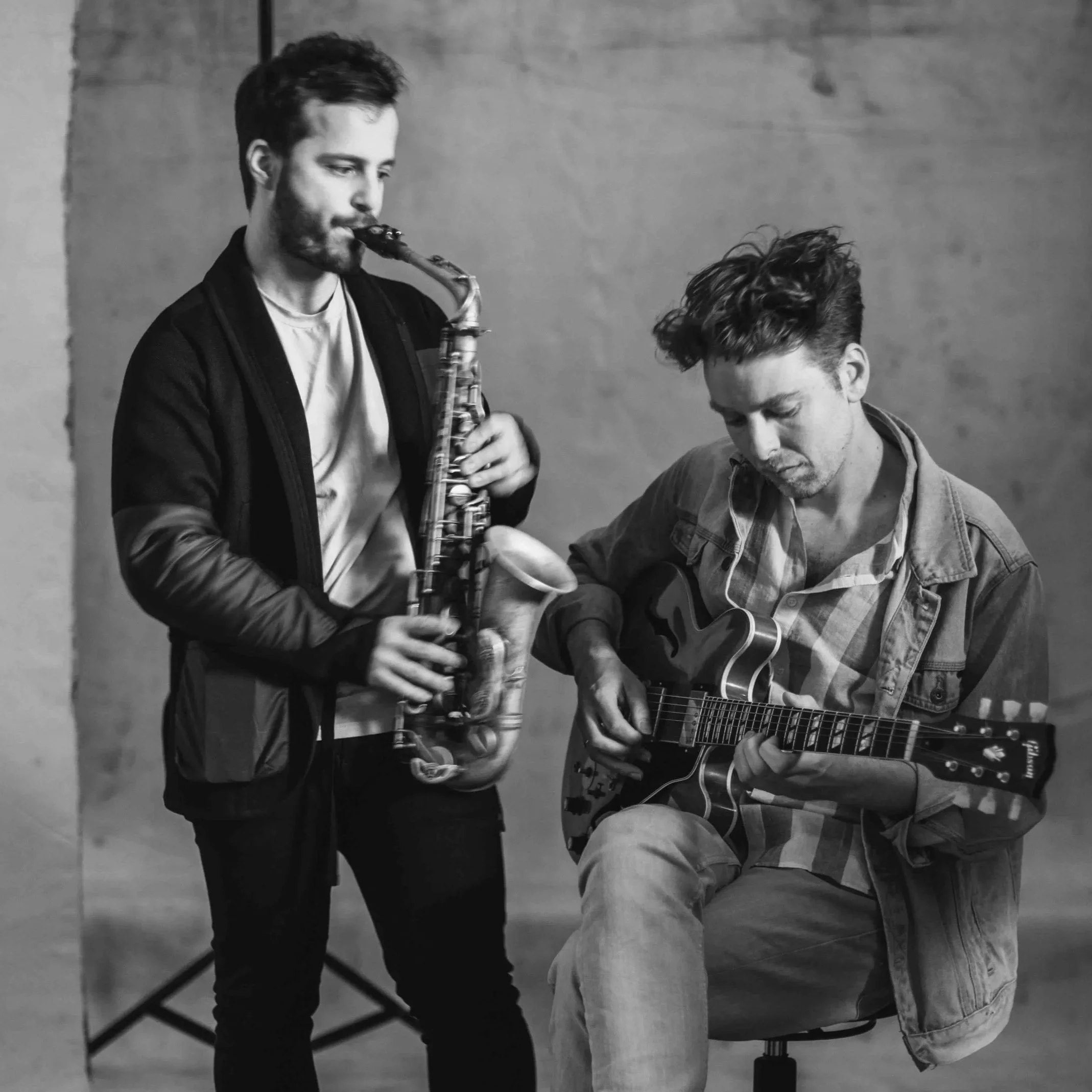 Two hip young men, one playing an acoustic guitar and the other playing a saxophone, in a minimalist studio setting with a plain wall background.