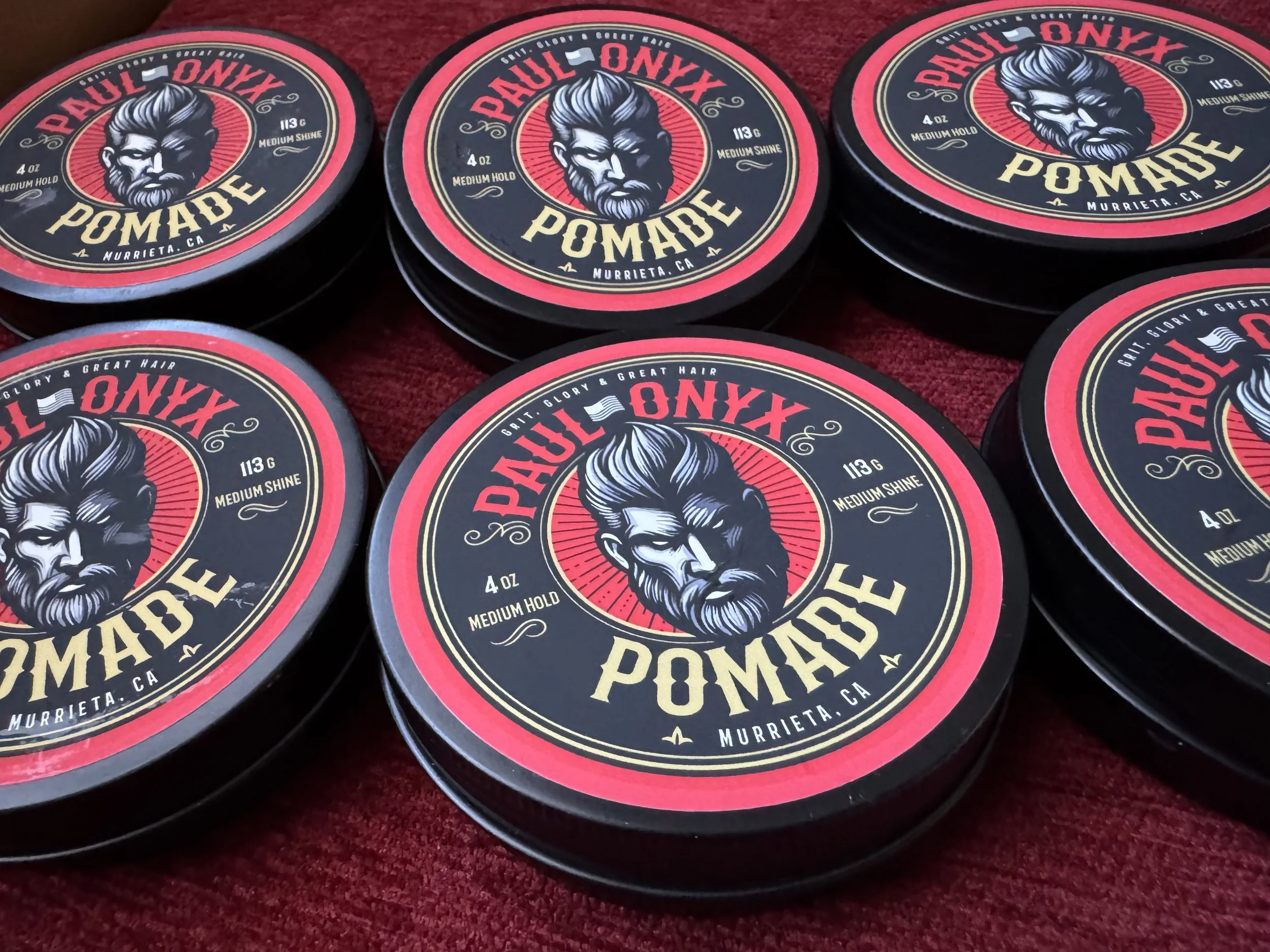 Multiple tins of Paul Ony Pomade with a vintage-style bearded man illustration on the label, placed on a red fabric surface.
