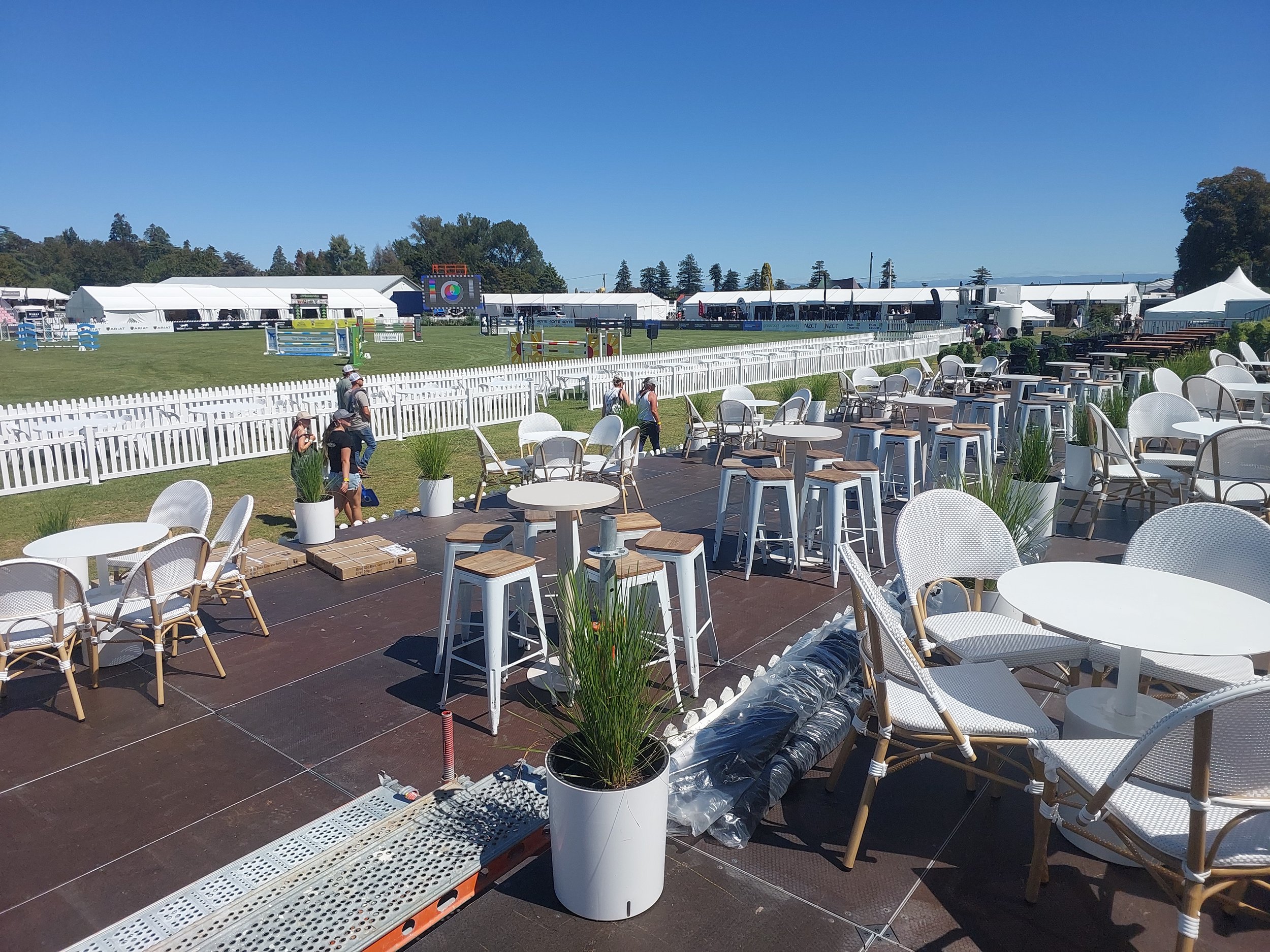 An outdoor scene of a horse show arena with a seating area in the foreground featuring white chairs, tables, and potted plants. The arena is fenced and several people are walking near the fence, with a large digital screen and tents in the background