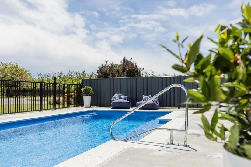 An outdoor swimming pool with blue water, a metal handrail, a black fence, and patio furniture including a bean bag and cushions, with a cloudy sky and plants in the background.