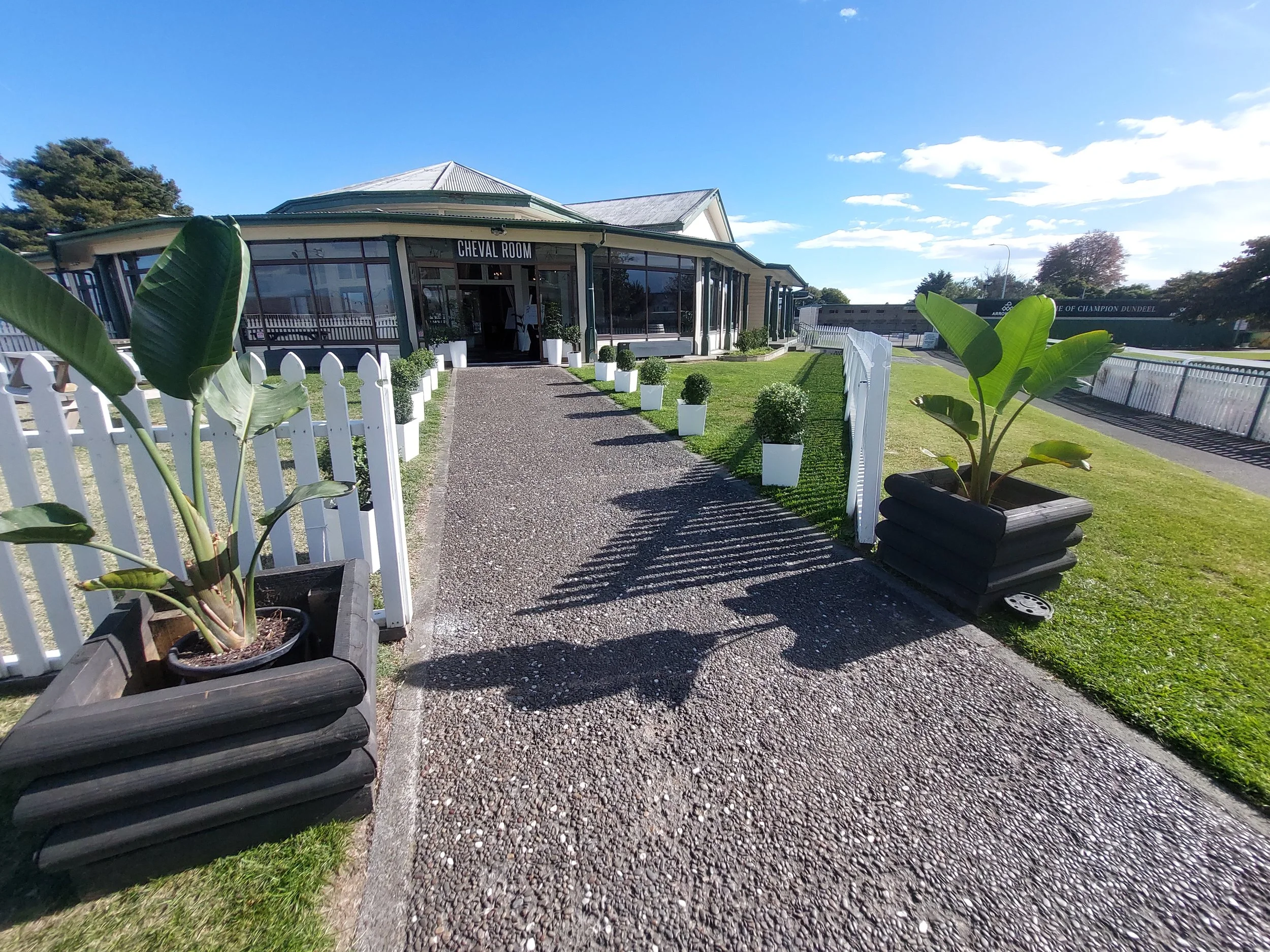 Pathway leading to a building labeled 'Cheval Room,' with potted plants on either side and a grassy area on the right, under a clear blue sky.
