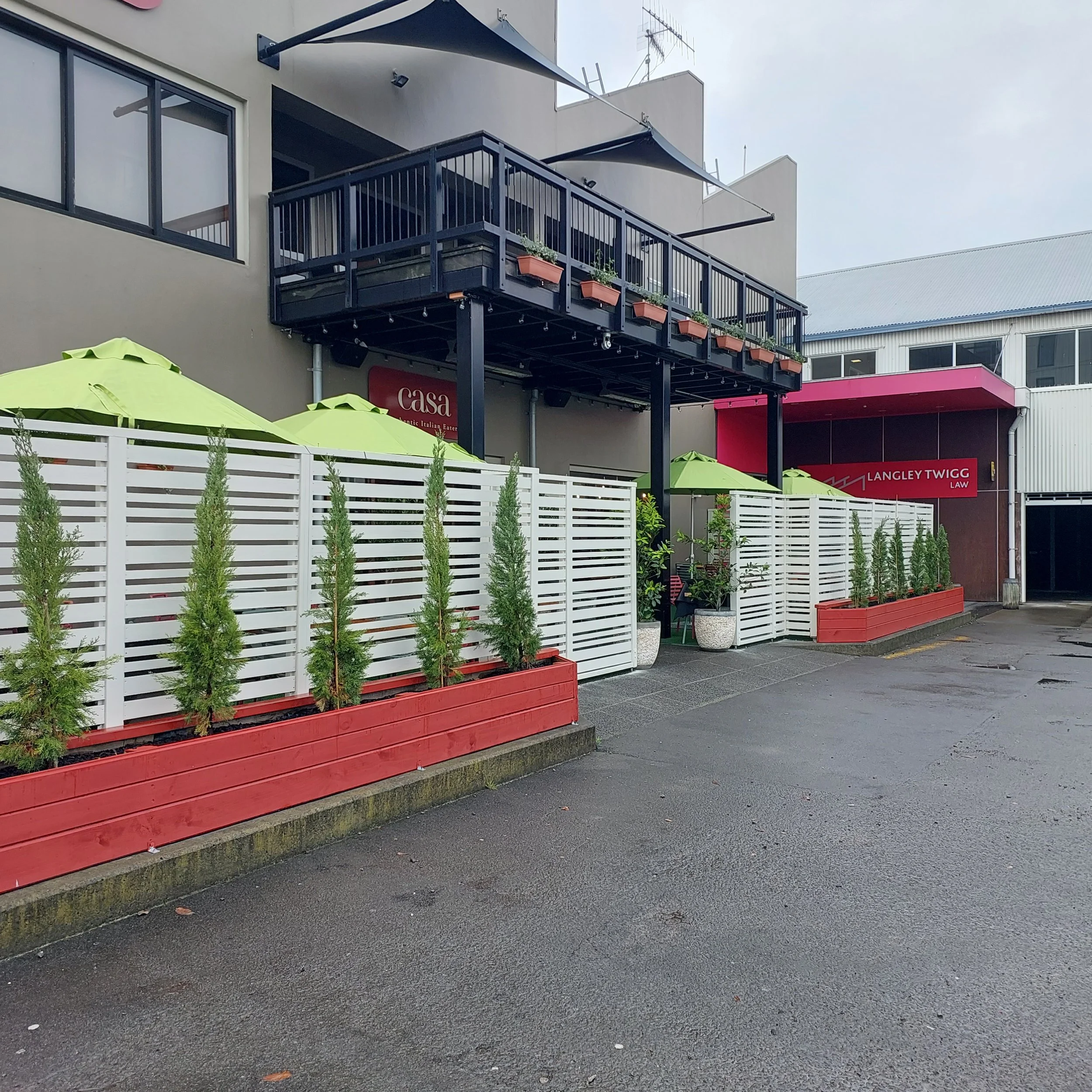Exterior view of a restaurant with a white fence, potted plants, green umbrellas, and a raised balcony with potted plants. Signage reads 'Casa' and 'Langley Twigg Law'.