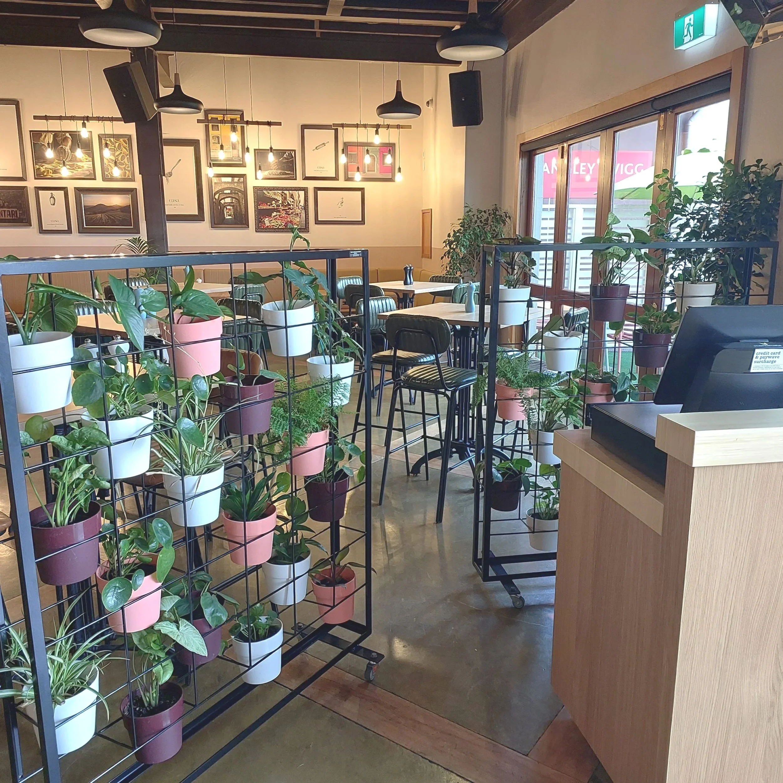 Interior of a cafe with seating area, decorated with hanging pendant lights and framed artwork on the wall. The space is separated by black metal plant stands filled with various potted plants.