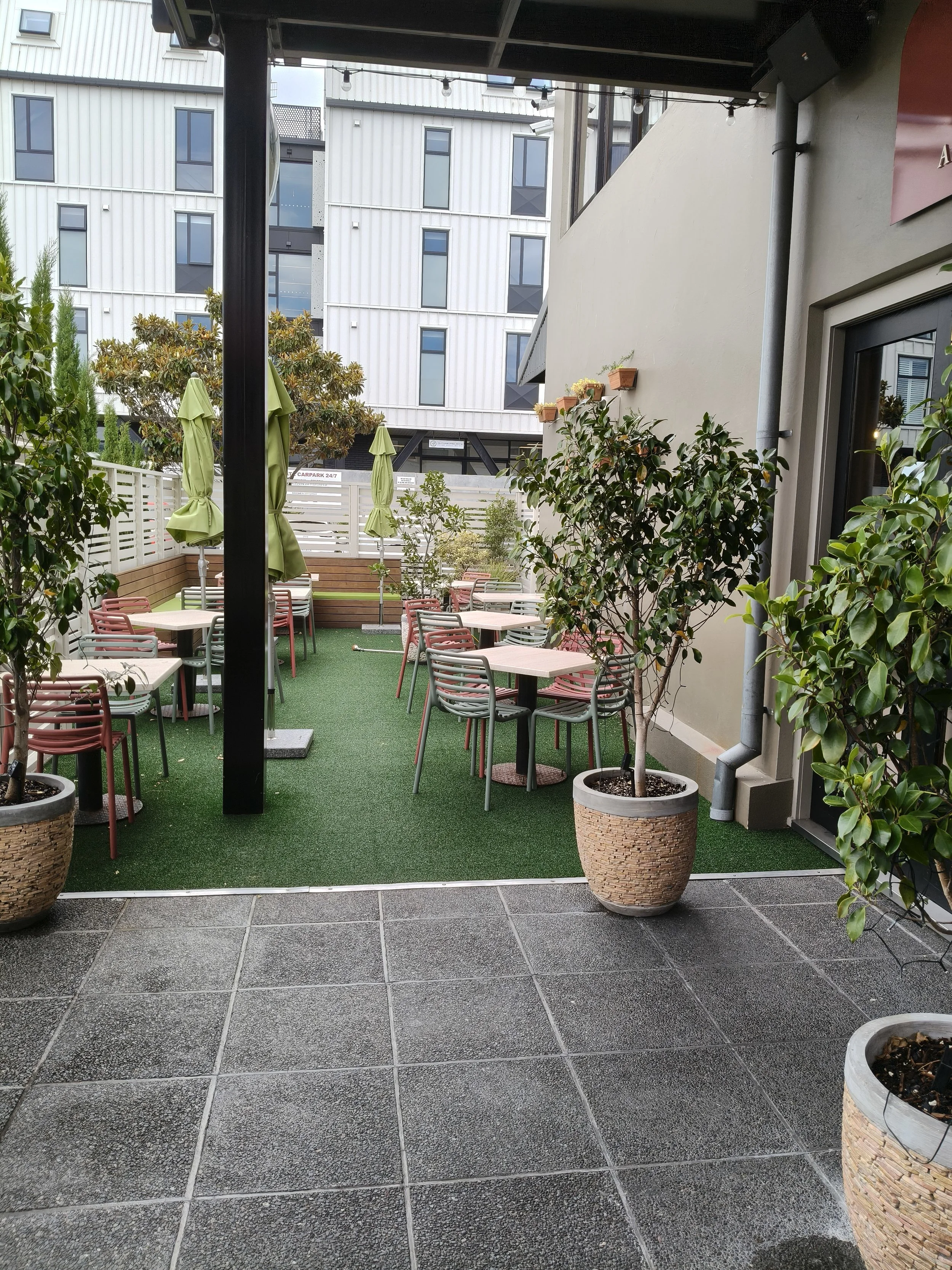 An outdoor patio with potted plants, several tables and chairs, green umbrellas, and a raised deck area in the background, surrounded by a white fence and modern buildings.