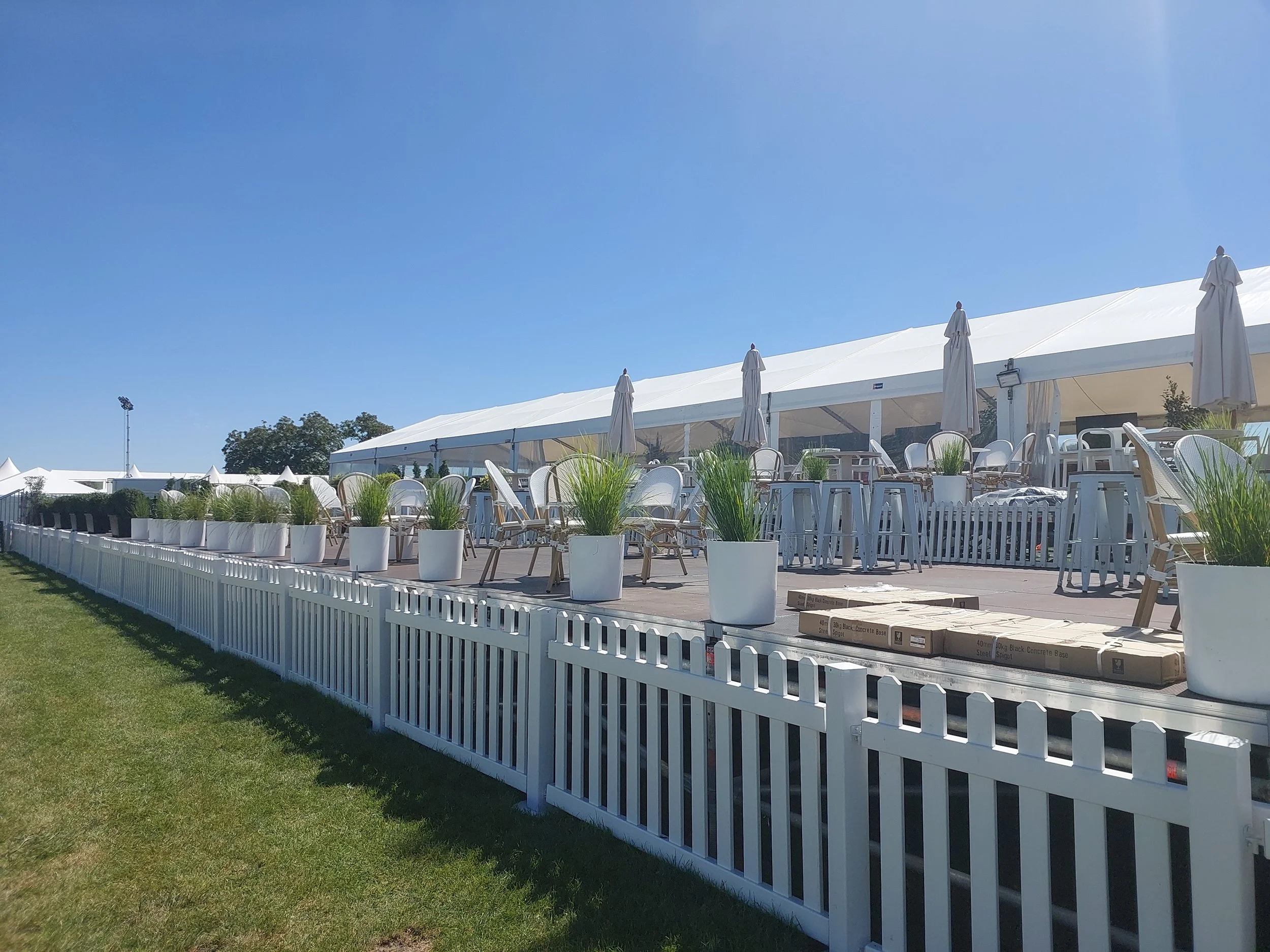 Empty outdoor event tent with white tables, chairs, umbrellas, and potted plants, surrounded by a white picket fence on a sunny day.