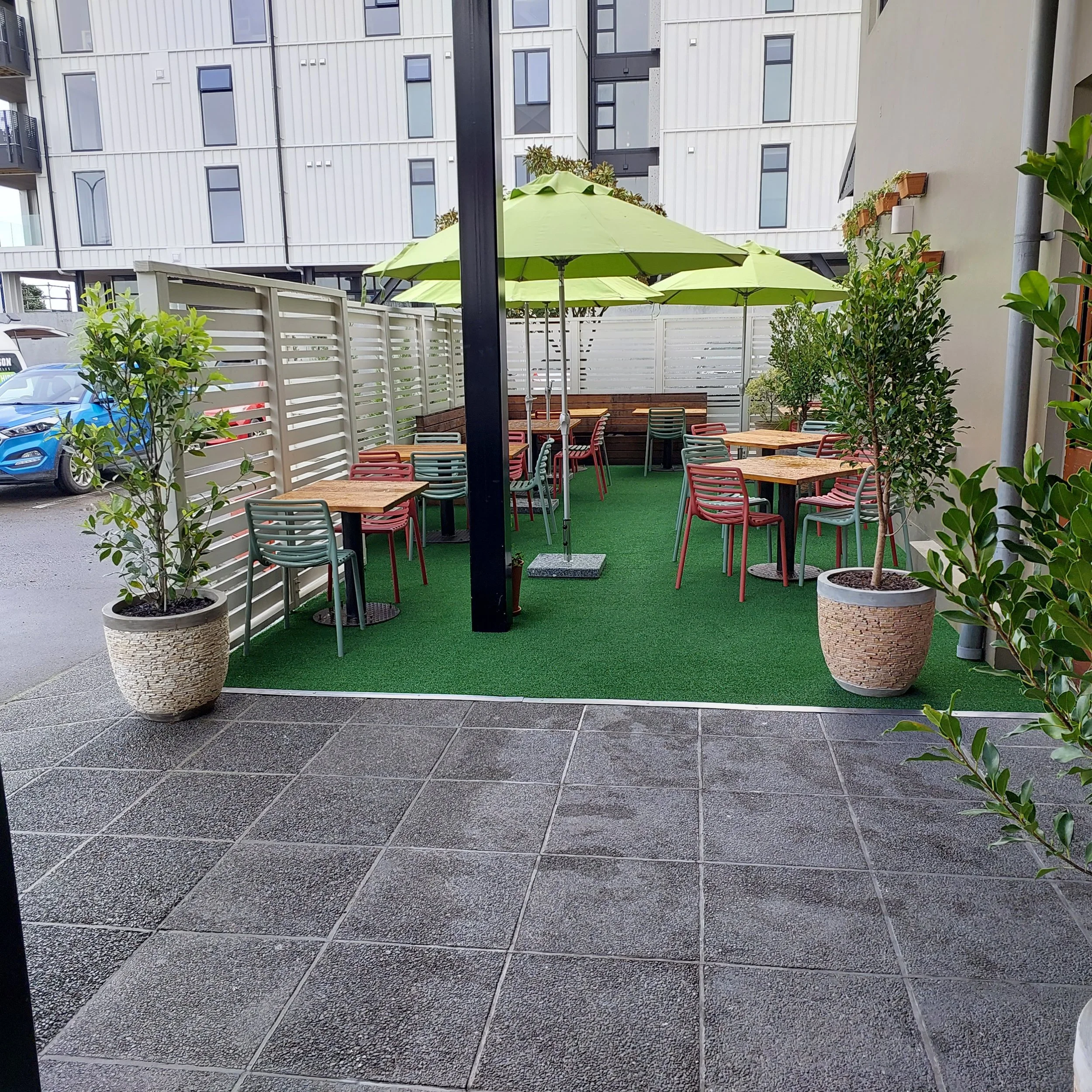 Outdoor patio area with several wooden tables and colorful chairs, two large green umbrellas, potted plants, and artificial grass flooring, enclosed by a white fence, with a residential building in the background.