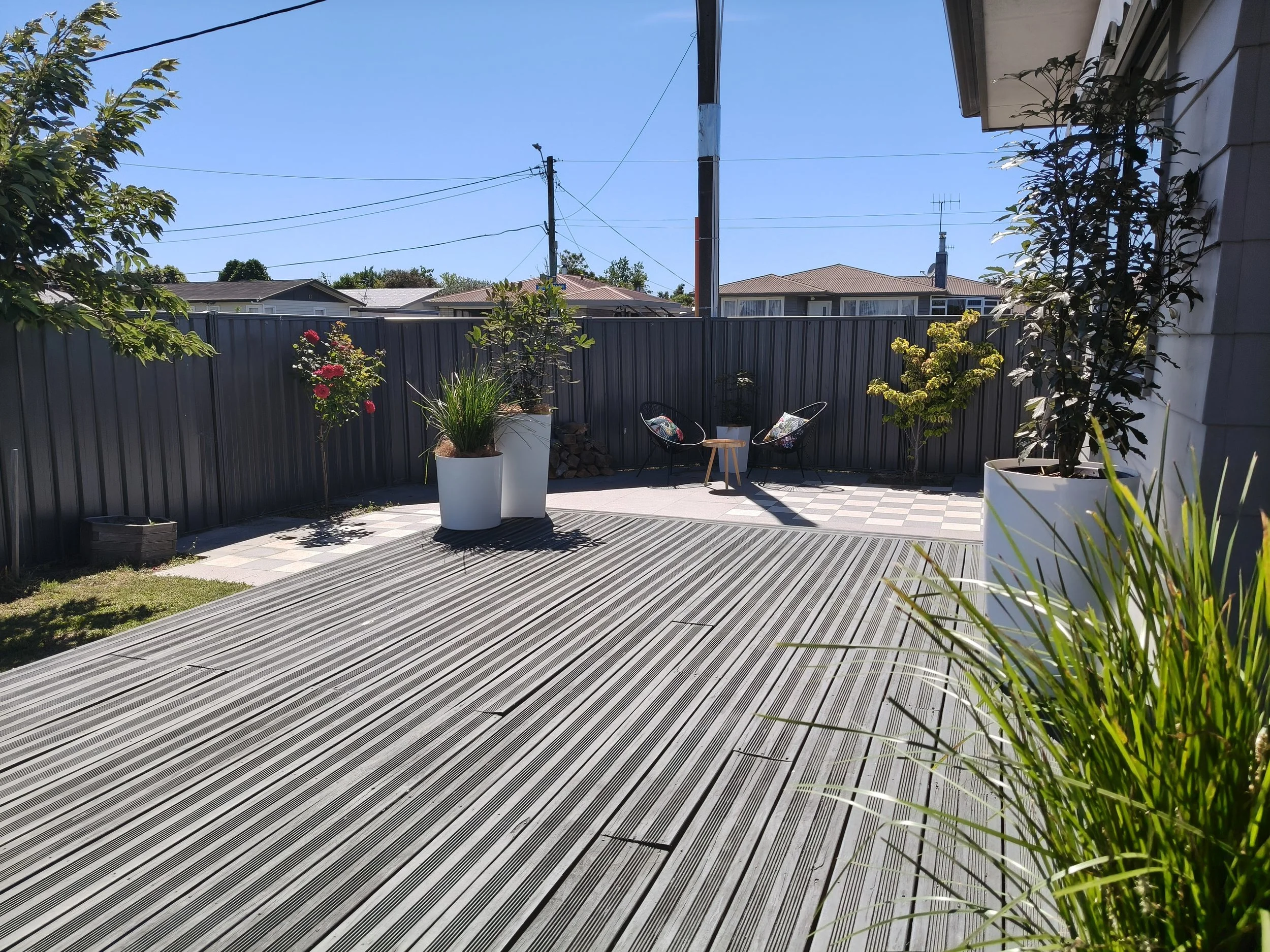 A backyard patio area with potted plants, two black chairs with floral cushions, a small wooden table, and a grey fence in the background on a sunny day.