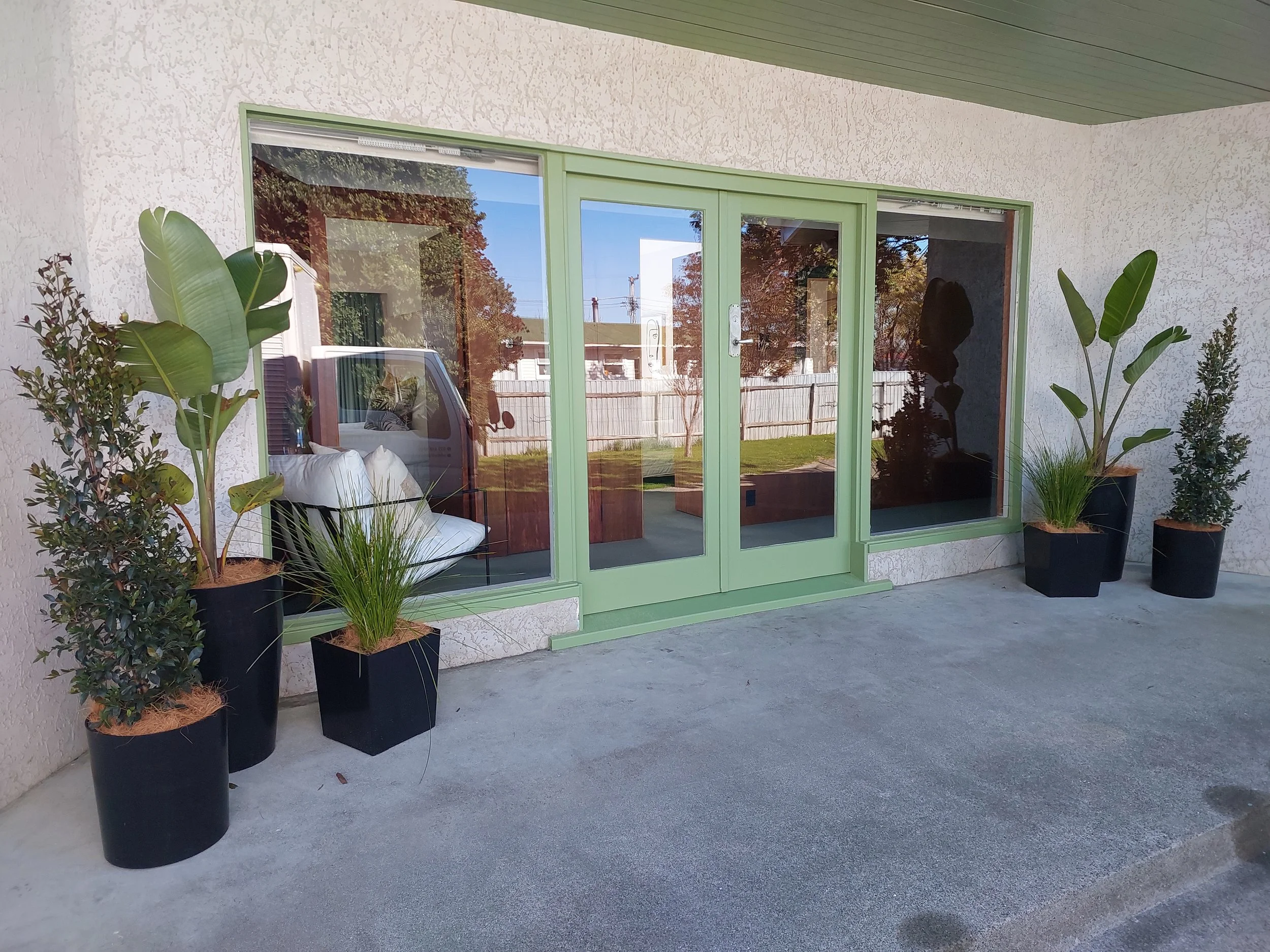 Exterior view of a house with large glass sliding doors framed in light green, surrounded by potted plants on a concrete patio.