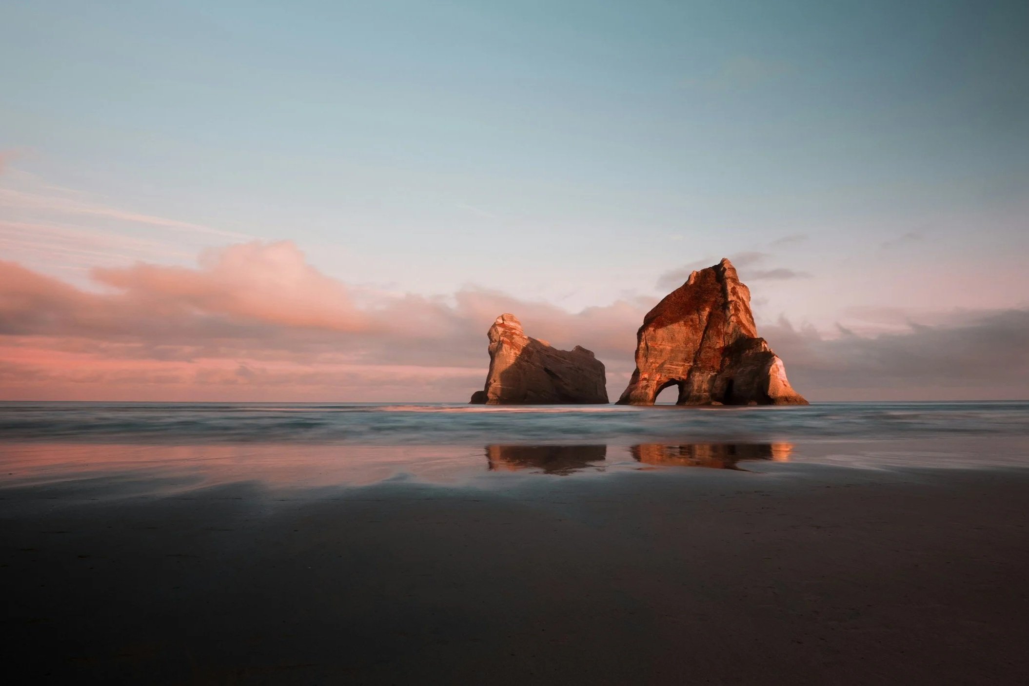 A beach with two large rock formations in the ocean, with one natural arch, during sunset with pink and orange clouds.