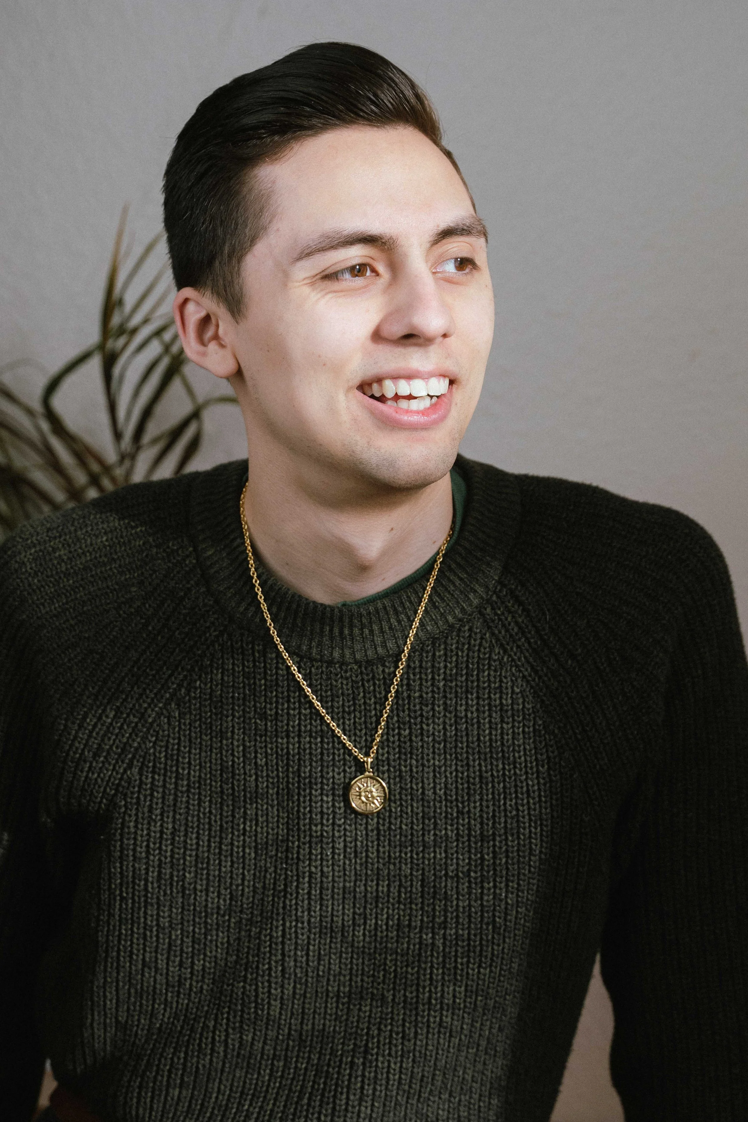Young man with dark hair smiling, wearing a black sweater and gold necklace, sitting in front of a neutral background with a plant in the background.