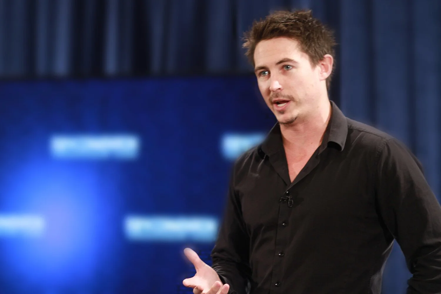 Man with dark hair, wearing a black shirt, speaking and gesturing with his hands in front of a blue curtain background.