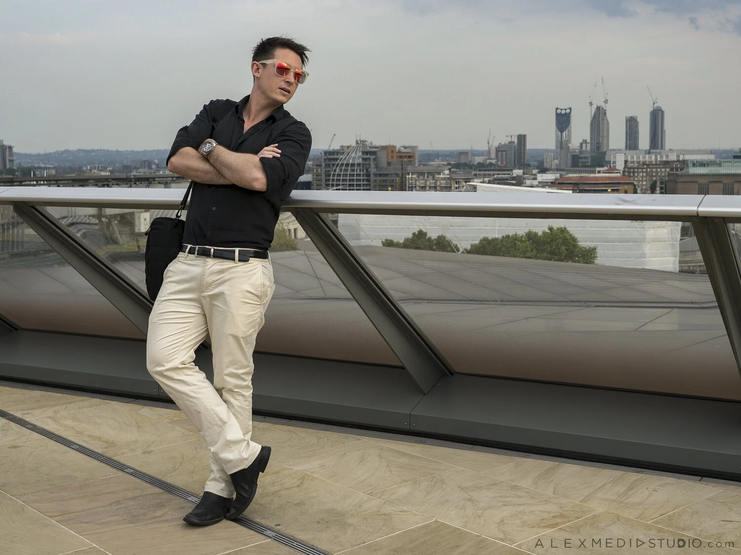 A man with sunglasses, black shirt, beige pants, and black shoes leaning against a glass railing on a rooftop, with the city skyline in the background.