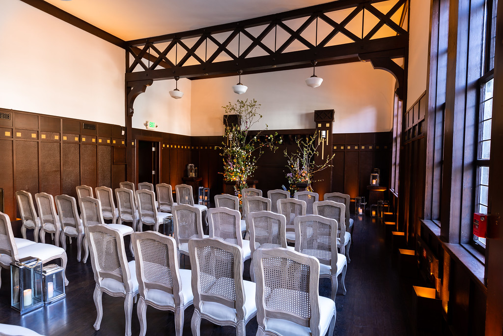 Indoor wedding or event venue with rows of white chairs, dark wood paneling, floral arrangements, and large windows on the right
