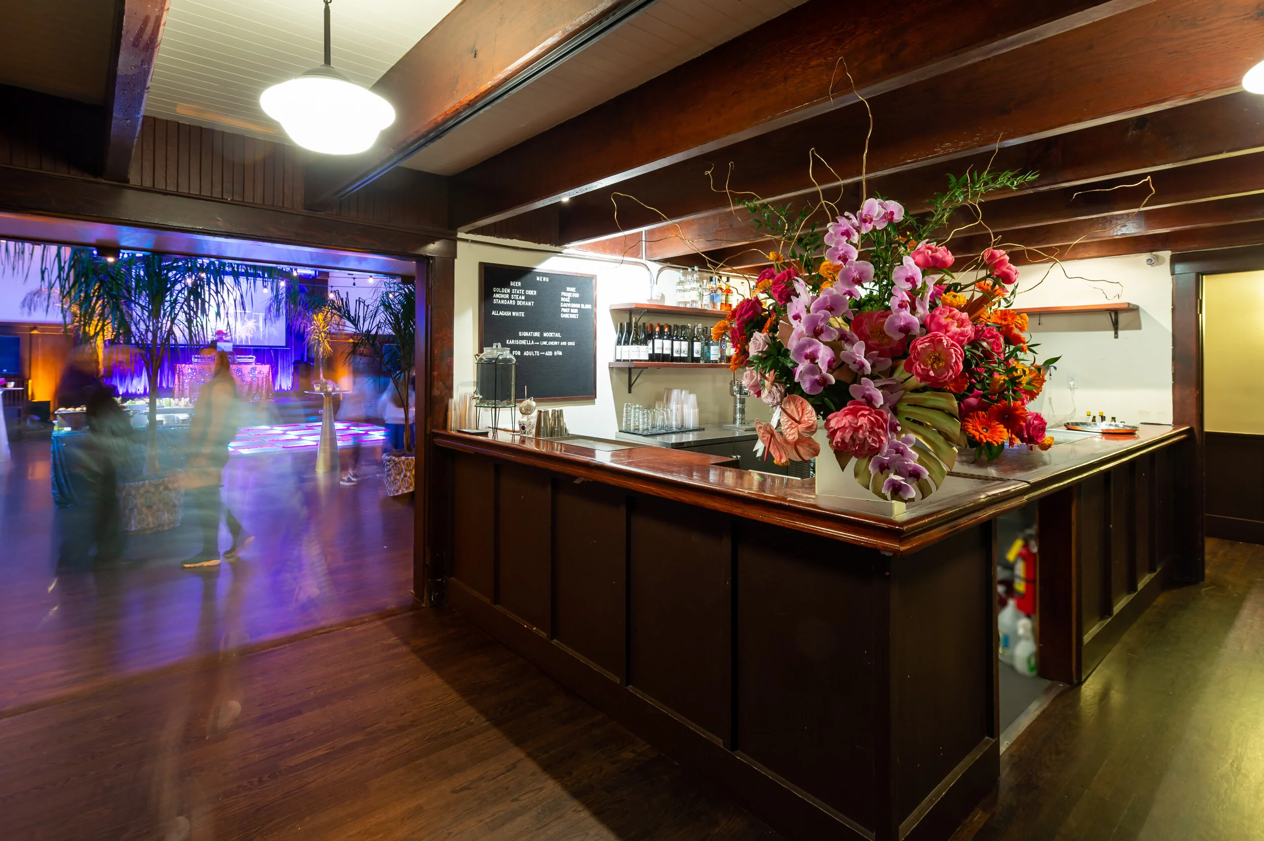 A dark wooden bar with a large colorful flower arrangement on the counter, in a cozy dimly lit venue with a dance floor and purple lighting in the background.