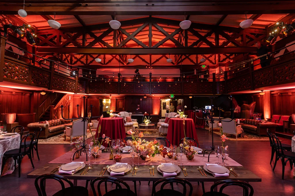 A warmly lit, elegantly decorated event space with a high wooden ceiling, round and rectangular tables with floral centerpieces, and a seating area with couches on a polished wooden floor.