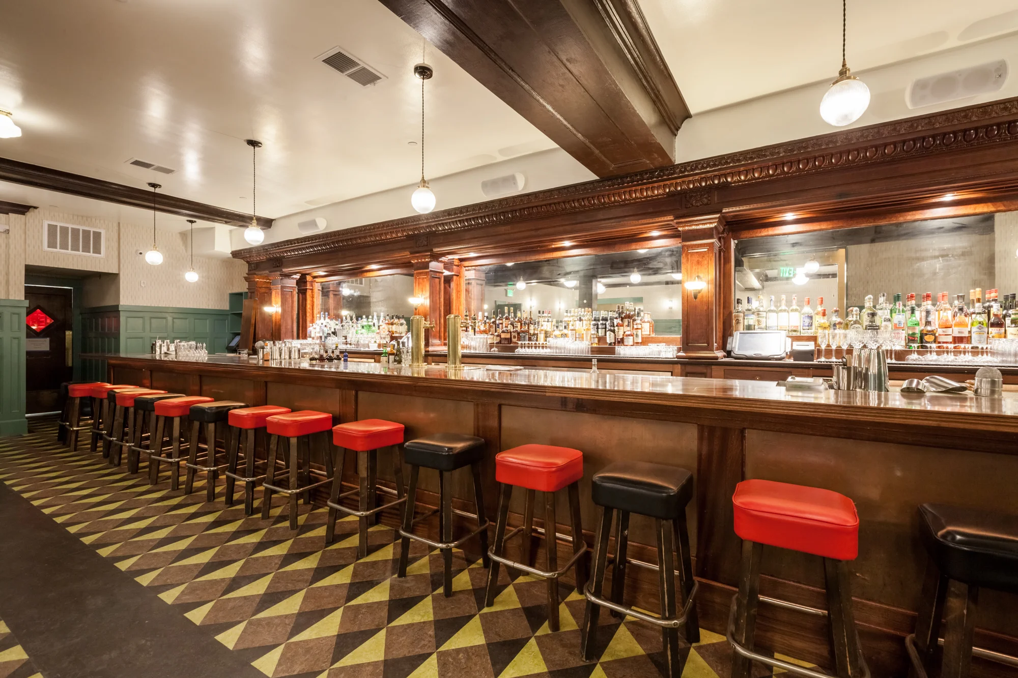 Empty bar with a wooden counter and red barstools in a dimly lit restaurant or bar interior. Shelves behind the counter are stocked with various bottles of alcohol and glasses.