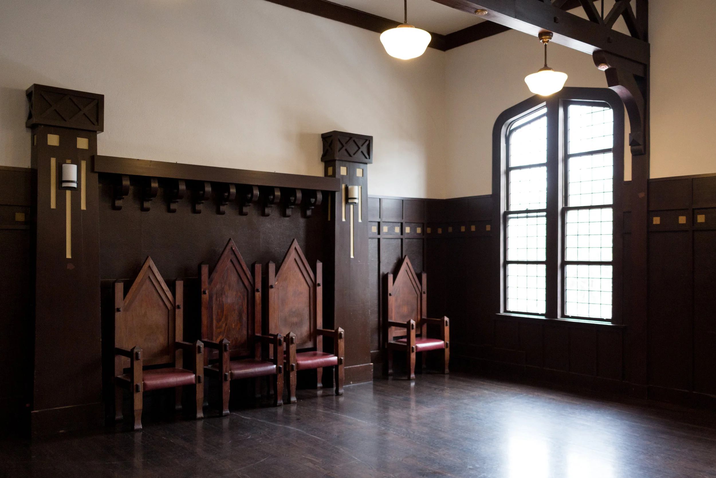 Empty room with dark wooden paneling, three chairs with tall spiked backs and red cushions, large arched window with grid pattern, and two ceiling lights.