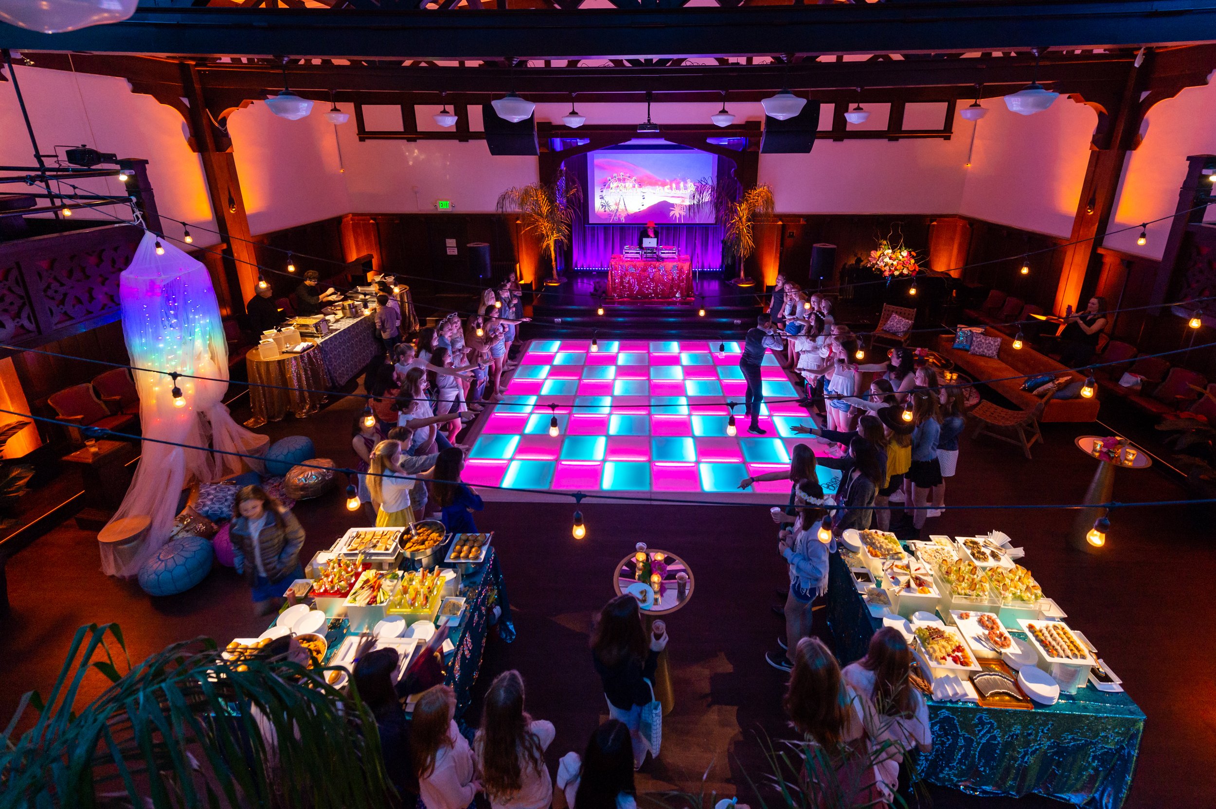 A large indoor party or dance event with a spacious illuminated dance floor, surrounded by guests, and a DJ booth at the back with a large screen displaying visuals. There are decorated tables with food and an area for seating, with warm lighting and