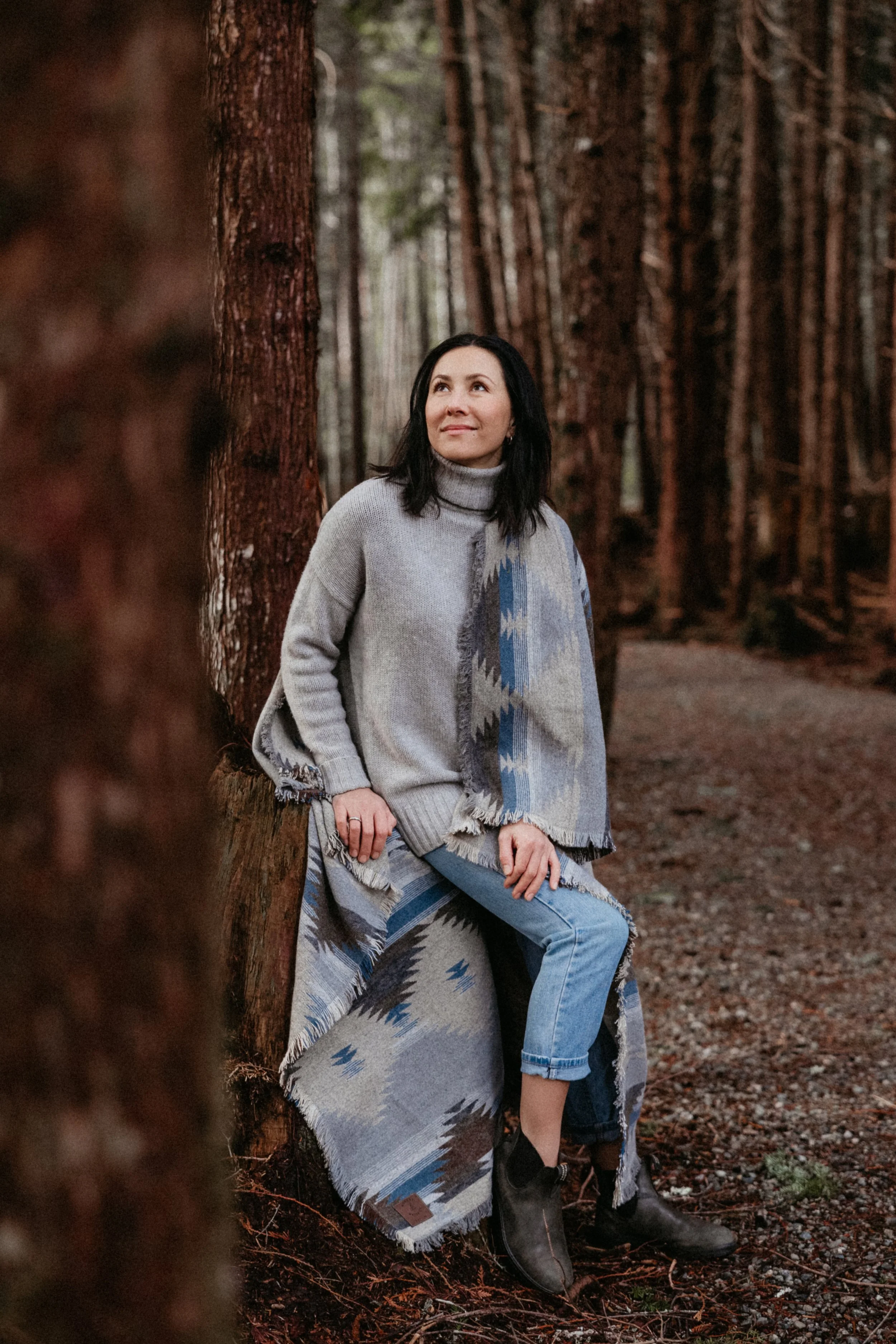 Woman in a gray turtleneck sweater and blue jeans, sitting on a tree trunk in a dense forest, looking upward, with a patterned blanket draped over her shoulders.