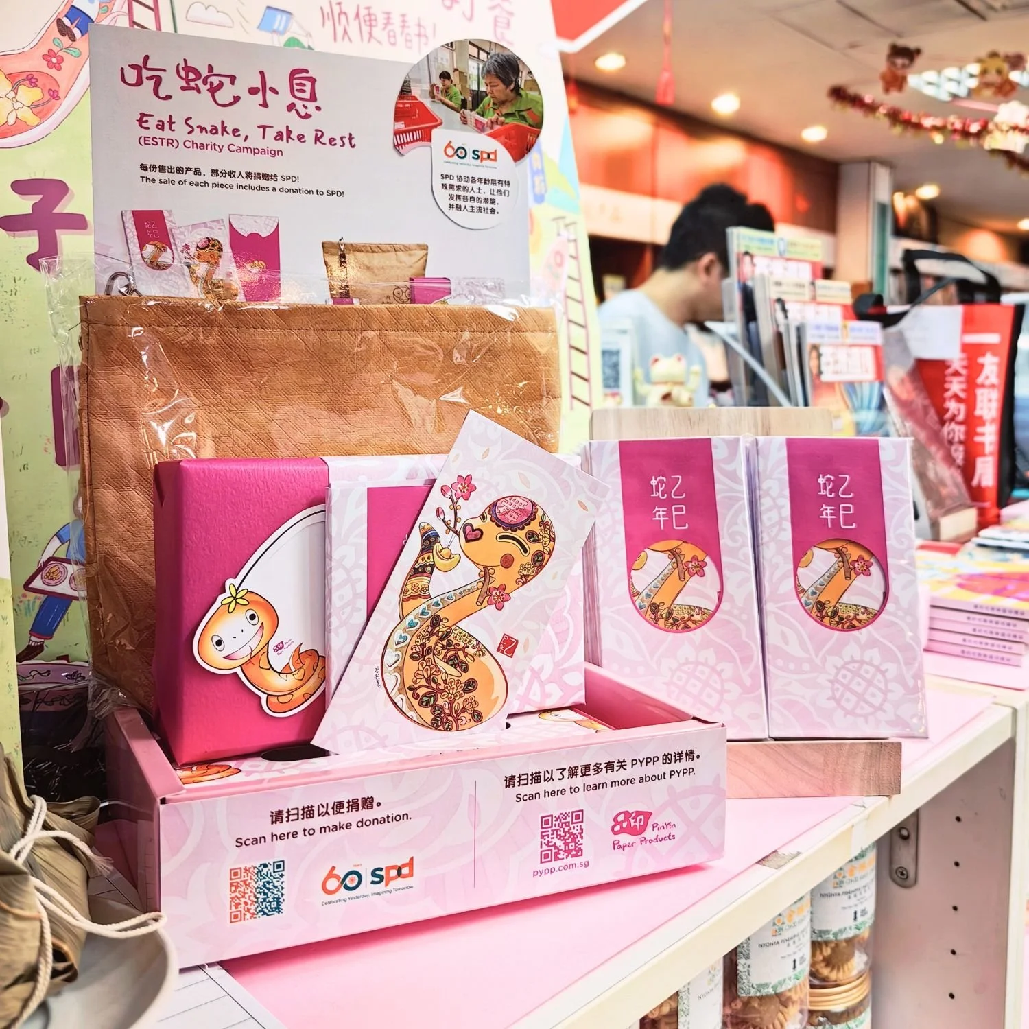 Retail counter display showcasing the pink Eat Snake Take Rest campaign gift packs and Hong Bao, informational panels, and donation QR codes for SPD.