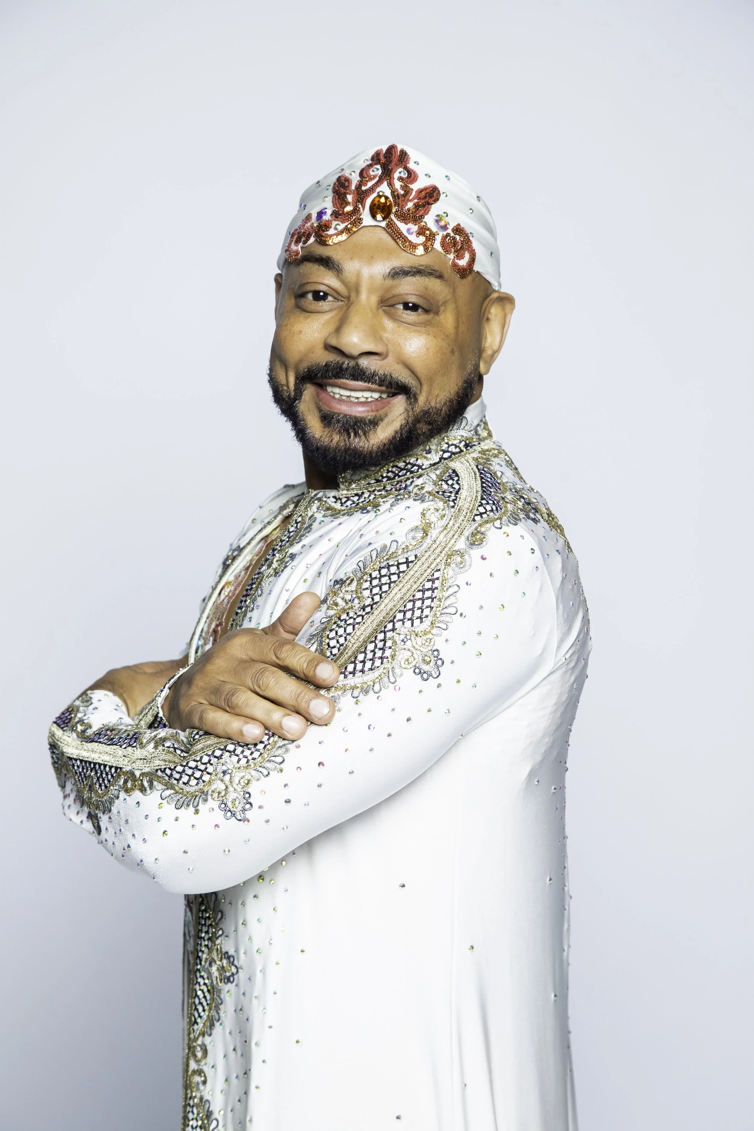 A man smiling, wearing a white ornate traditional outfit with intricate embroidery and embellishments, and a matching head covering.