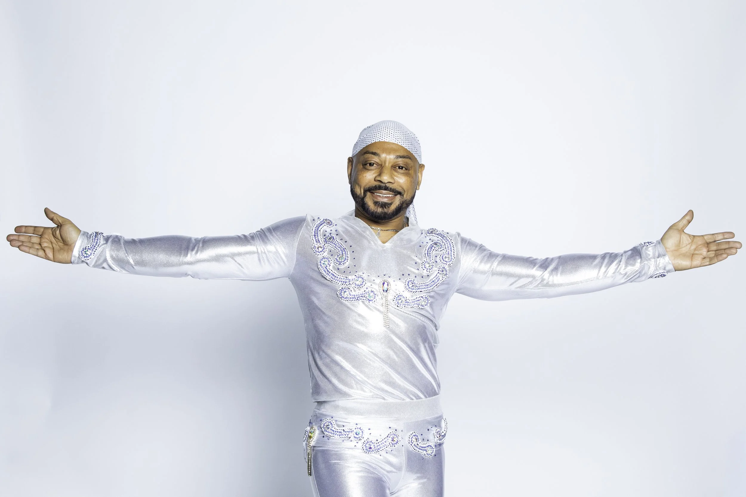Man dressed in a shiny silver outfit with embroidered details, wearing a matching cap, extending his arms, smiling, against a plain white background.