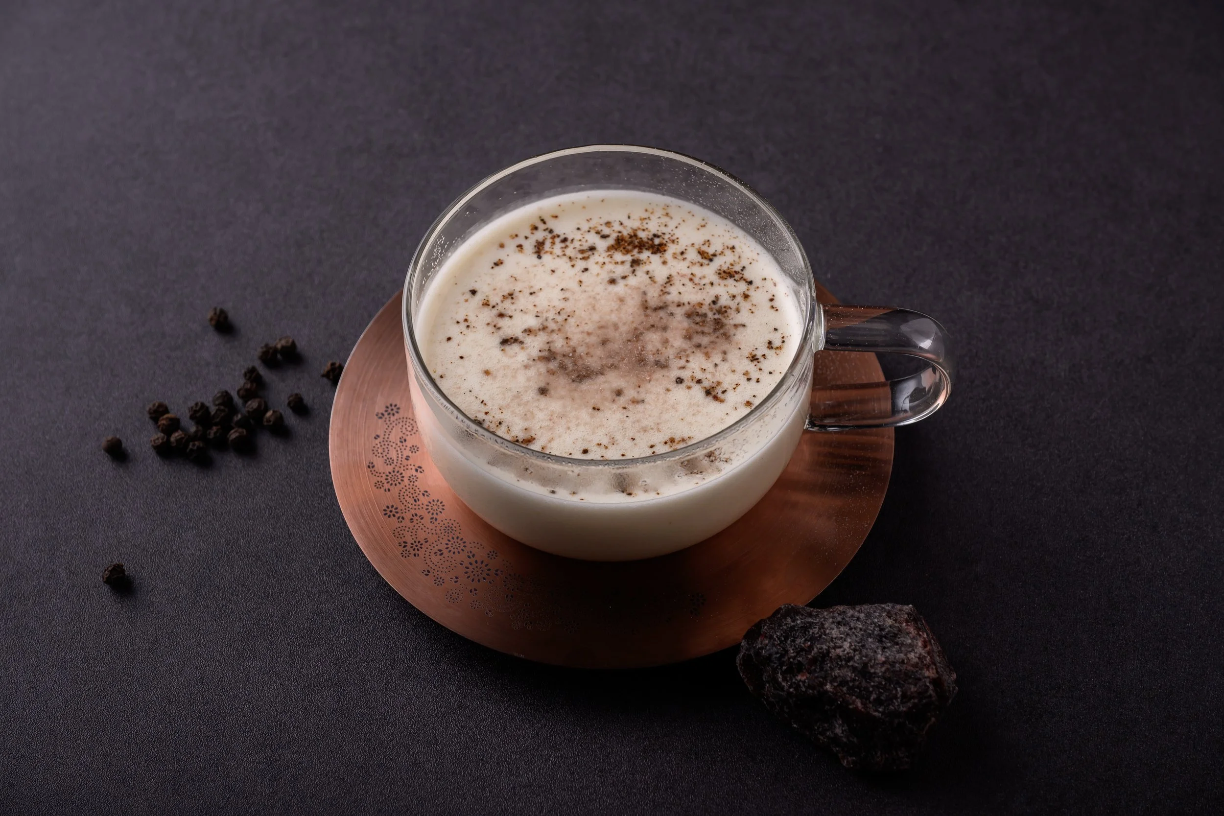 A cup of creamy beverage topped with ground spices, placed on a copper coaster with scattered black peppercorns and a piece of chocolate on a dark surface.