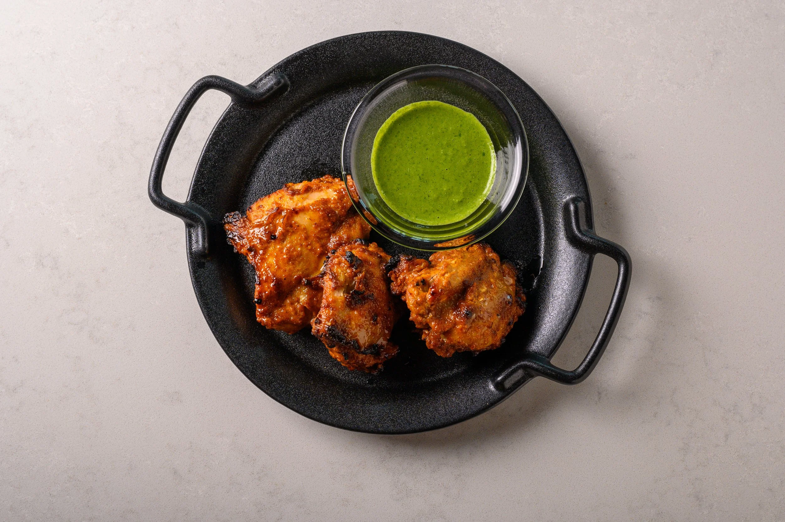 Three pieces of fried chicken with a side of green sauce in a small glass container on a black serving dish