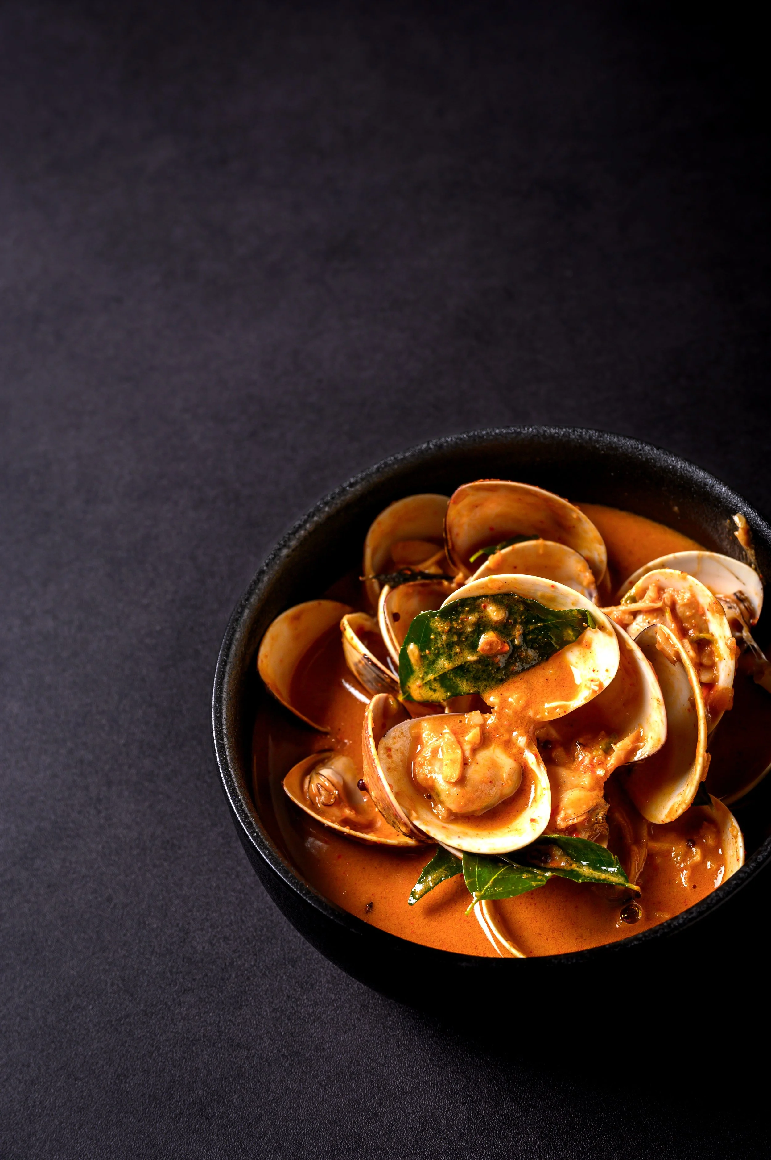 A bowl of clam soup with green herbs in a black bowl on a black surface.