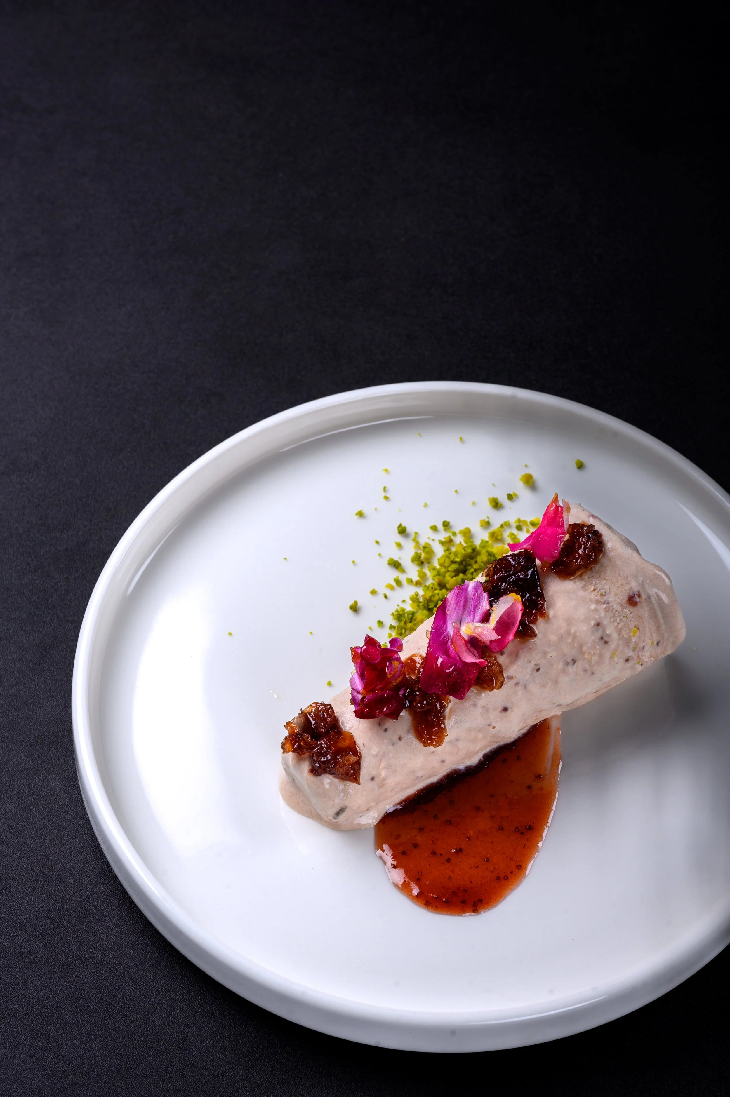 Dessert on a white plate with pink flower petals, crushed pistachios, and a caramel sauce