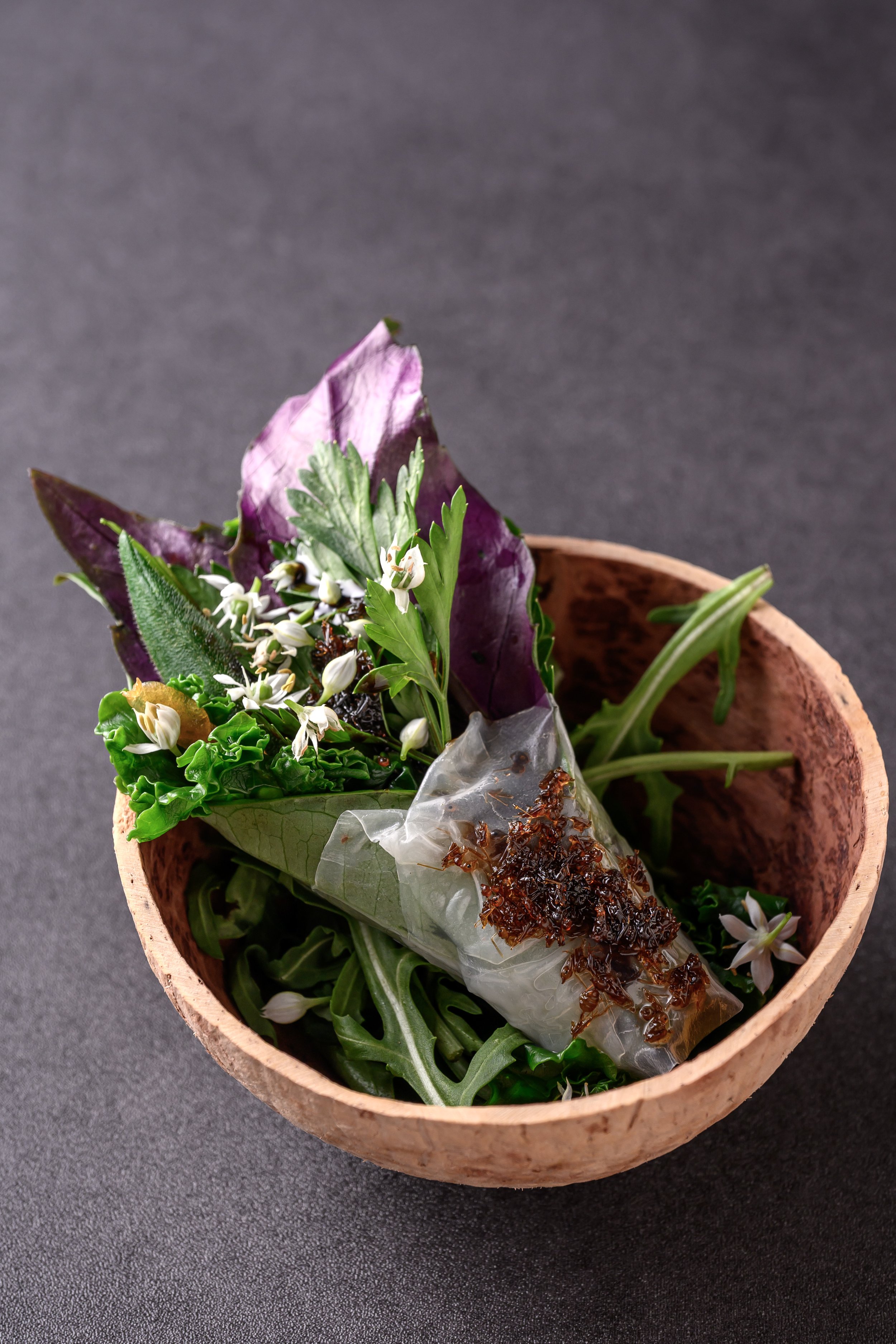 A wooden bowl with assorted fresh herbs, flowers, and a small glass container of sauce on a dark surface.