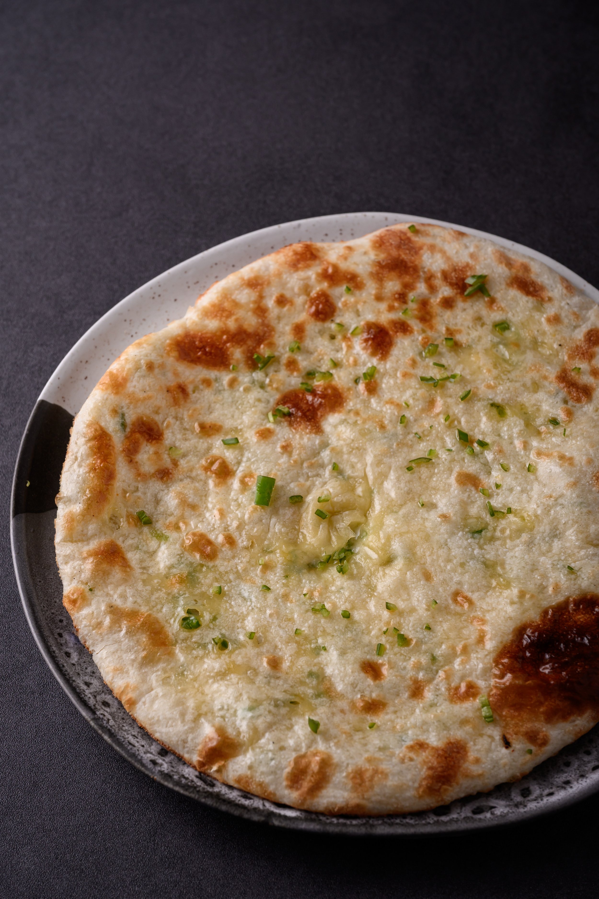 A close-up of a cheese pizza with green herbs on top, in a black skillet on a dark surface.
