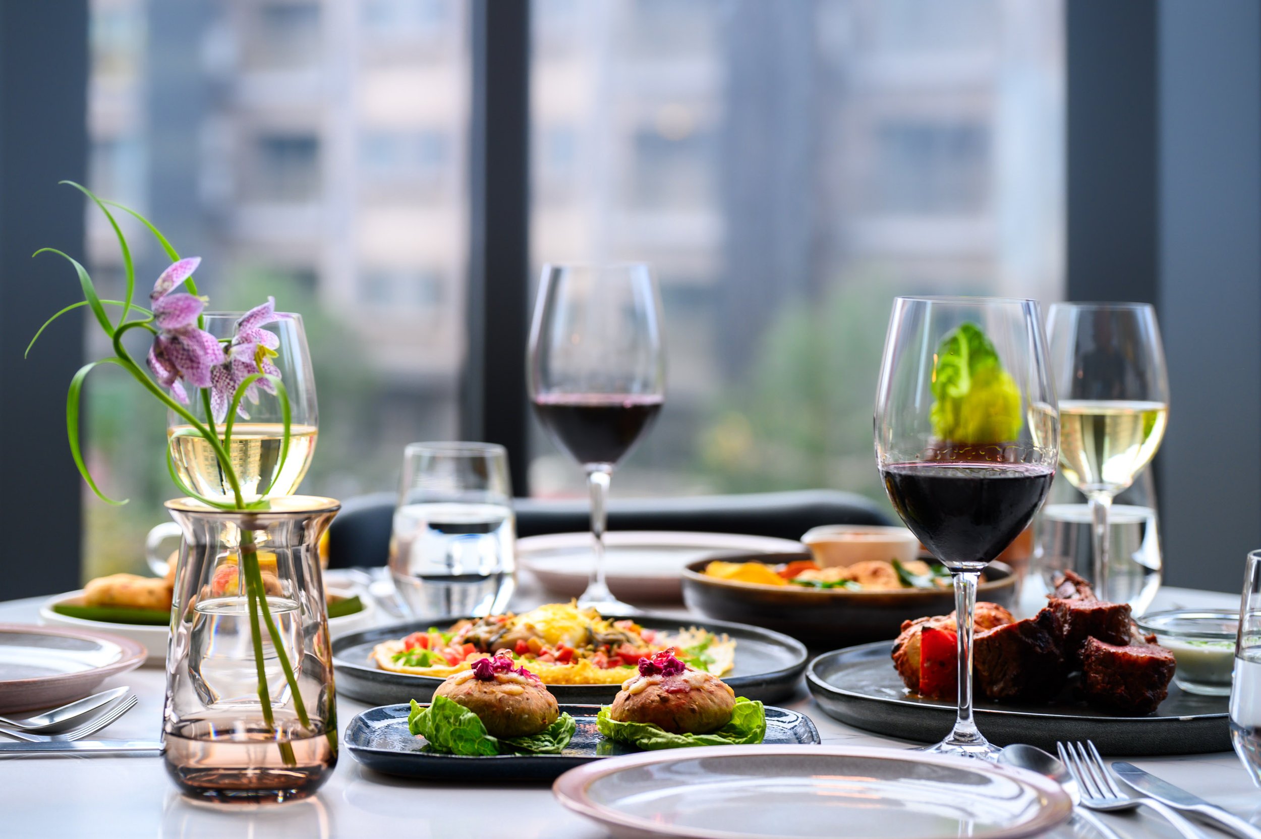 A dining table set with various dishes, glasses of red and white wine, water, a vase with purple flowers, and a city skyline view through large windows in the background.