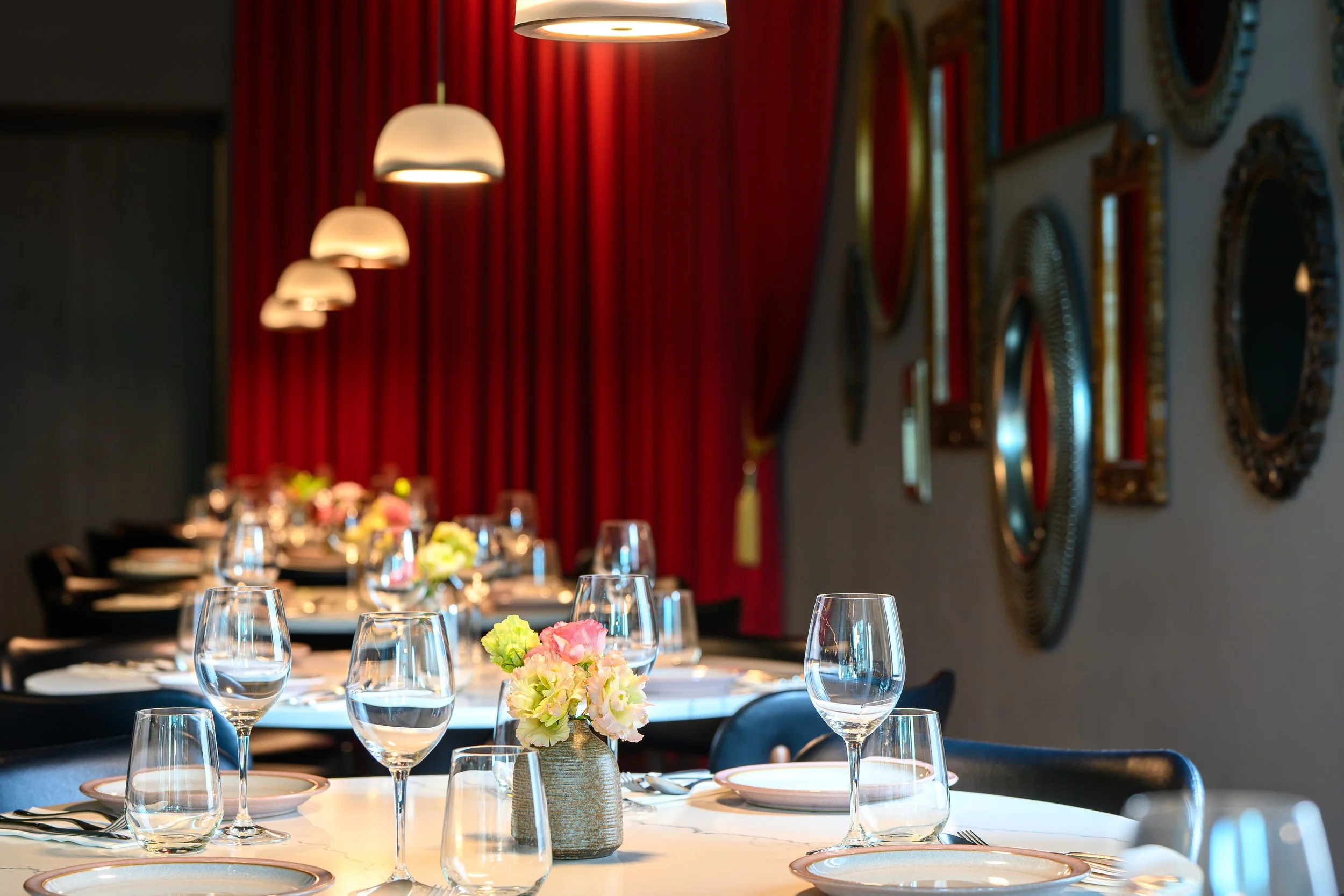 Elegant dining table set with glasses, plates, and a flower centerpiece in a restaurant with red curtains and mirrors on the wall.