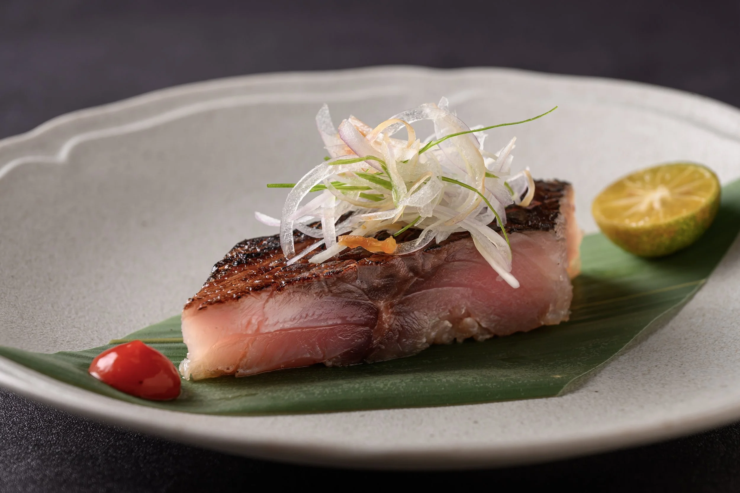 A piece of seared tuna with sliced onions and garnishes, served with a halved lime and a red cherry tomato on a green leaf on a white plate.