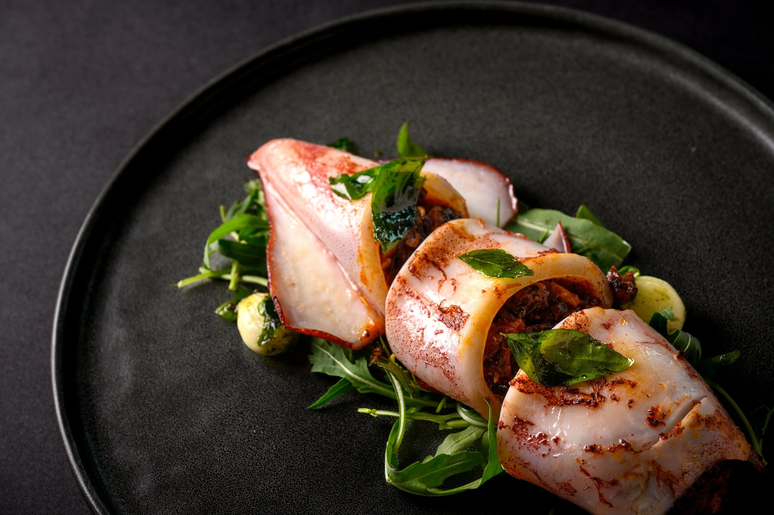 Grilled squid stuffed with a dark filling, served on a bed of fresh greens with basil leaves, on a black plate.
