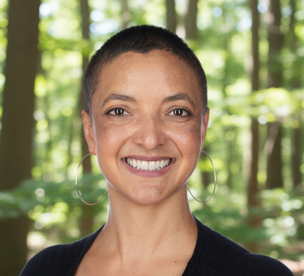 A woman with a short haircut smiling outdoors in a forest setting, wearing hoop earrings and a black top.