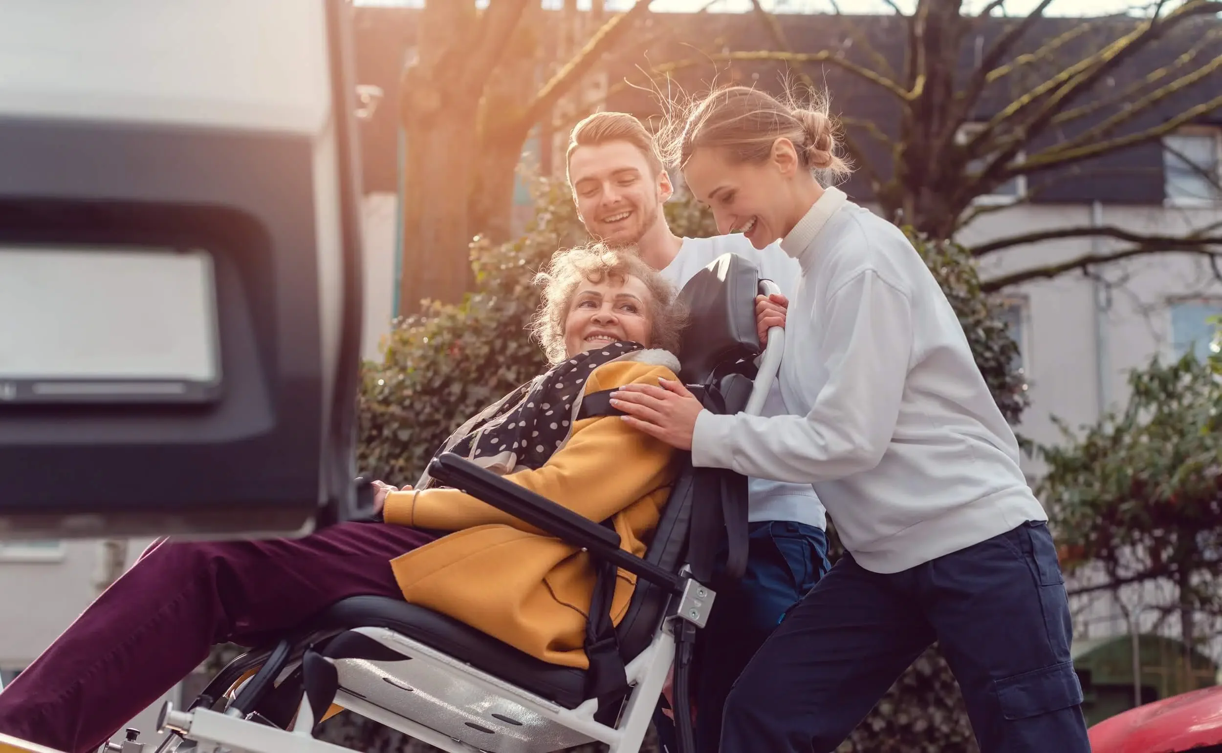 A caregiver pushing an elderly woman in a wheelchair while two young adults look at and smile at her outdoors during daytime.