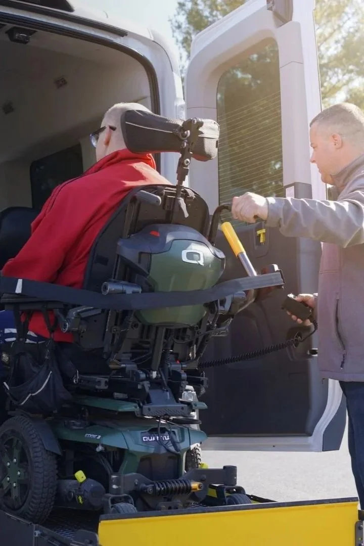 A person in a wheelchair being assisted into a van by another person holding a remote control. The wheelchair is equipped for mobility and the scene appears to be outdoors.