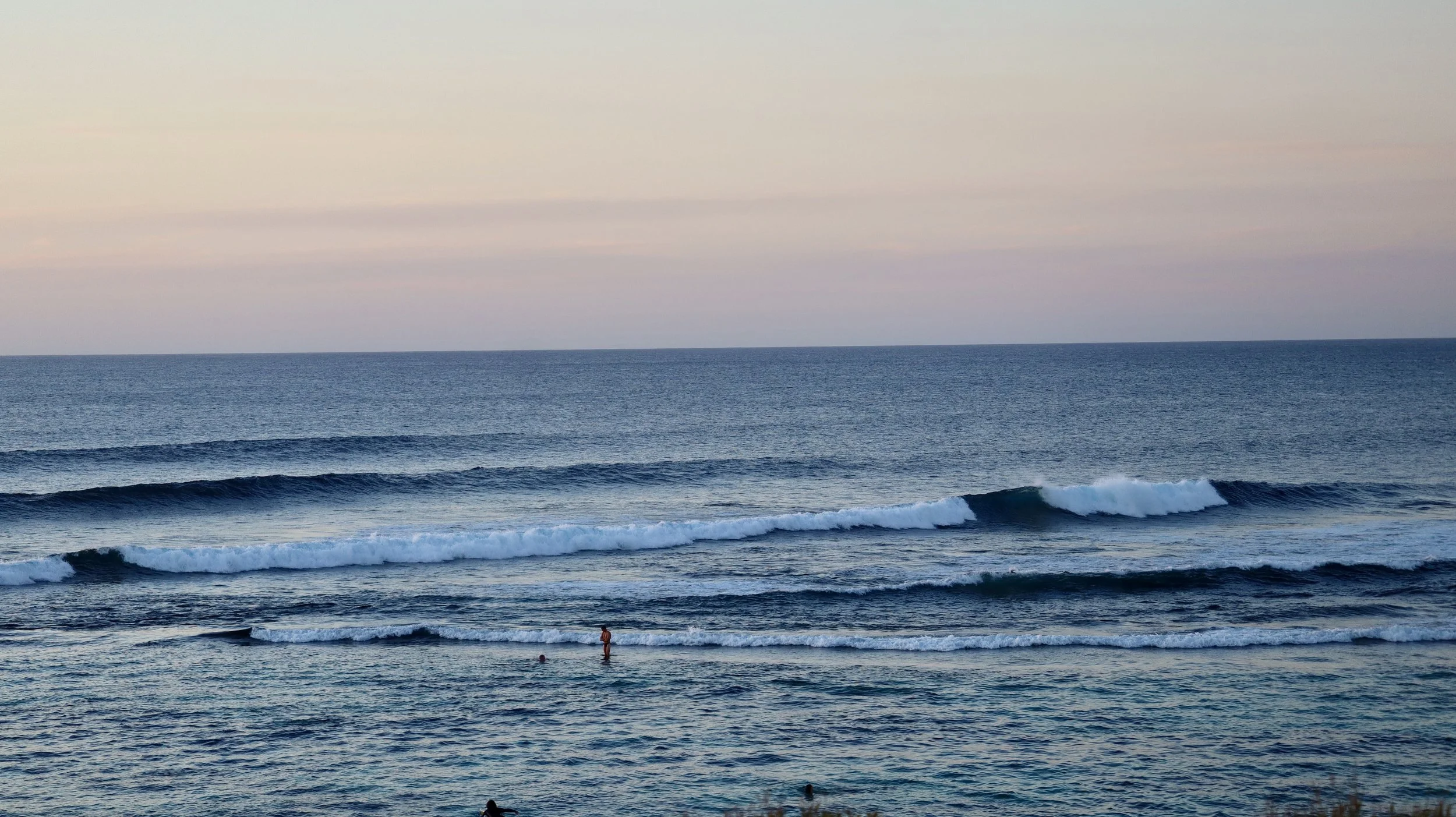 A photo of a calm Yallingyup ocean with small waves, a couple of people swimming and standing in the water, and a pastel-colored sky at sunset or sunrise, taken by Managing Director Jack Georgiu