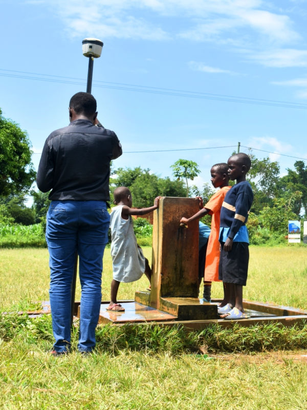 A man is taking a photo of children drawing water from a well in a grassy field with trees and sky in the background.