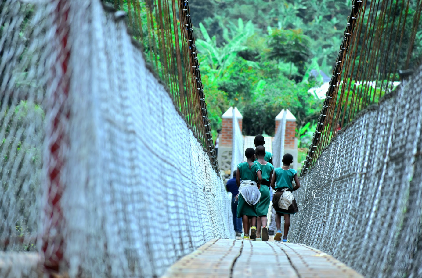 Children in school uniforms walk across a bridge over a lush green landscape.
